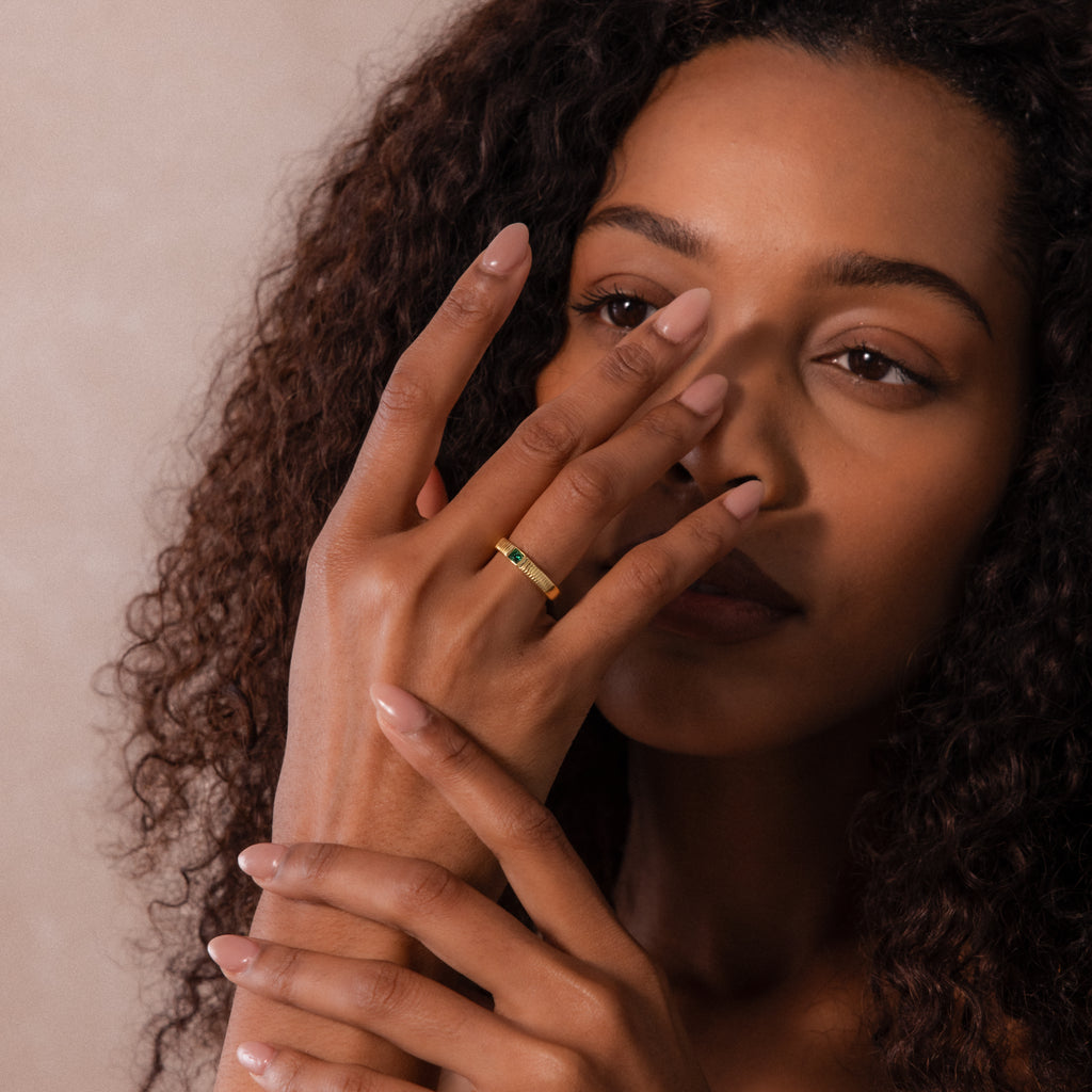A woman with curly hair touches her face, showcasing the Ribbed Emerald Band—a gold ring featuring a green square-cut stone—on her finger.