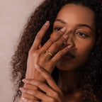 A woman with curly hair touches her face, showcasing the Ribbed Emerald Band—a gold ring featuring a green square-cut stone—on her finger.