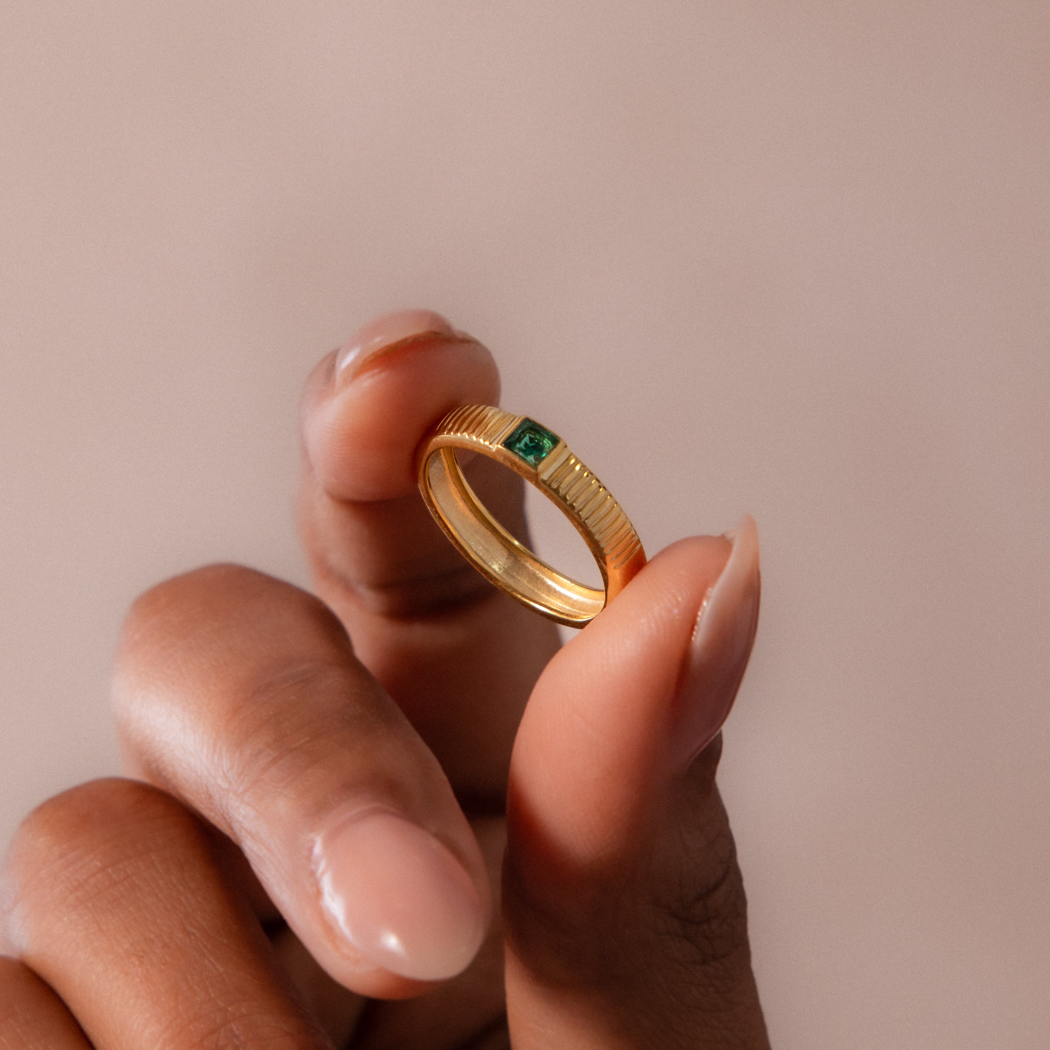 A hand holds the Ribbed Emerald Band, a gold birthstone ring featuring a green gemstone, set against a neutral background.