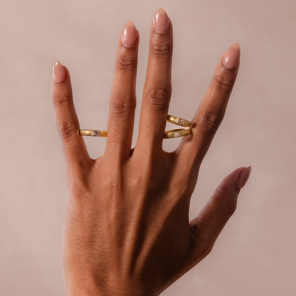 A hand with nude nails wears the Ribbed Peridot Band, a modern gold ring, on the middle finger and a square birthstone ring on the ring finger, set against a neutral background.