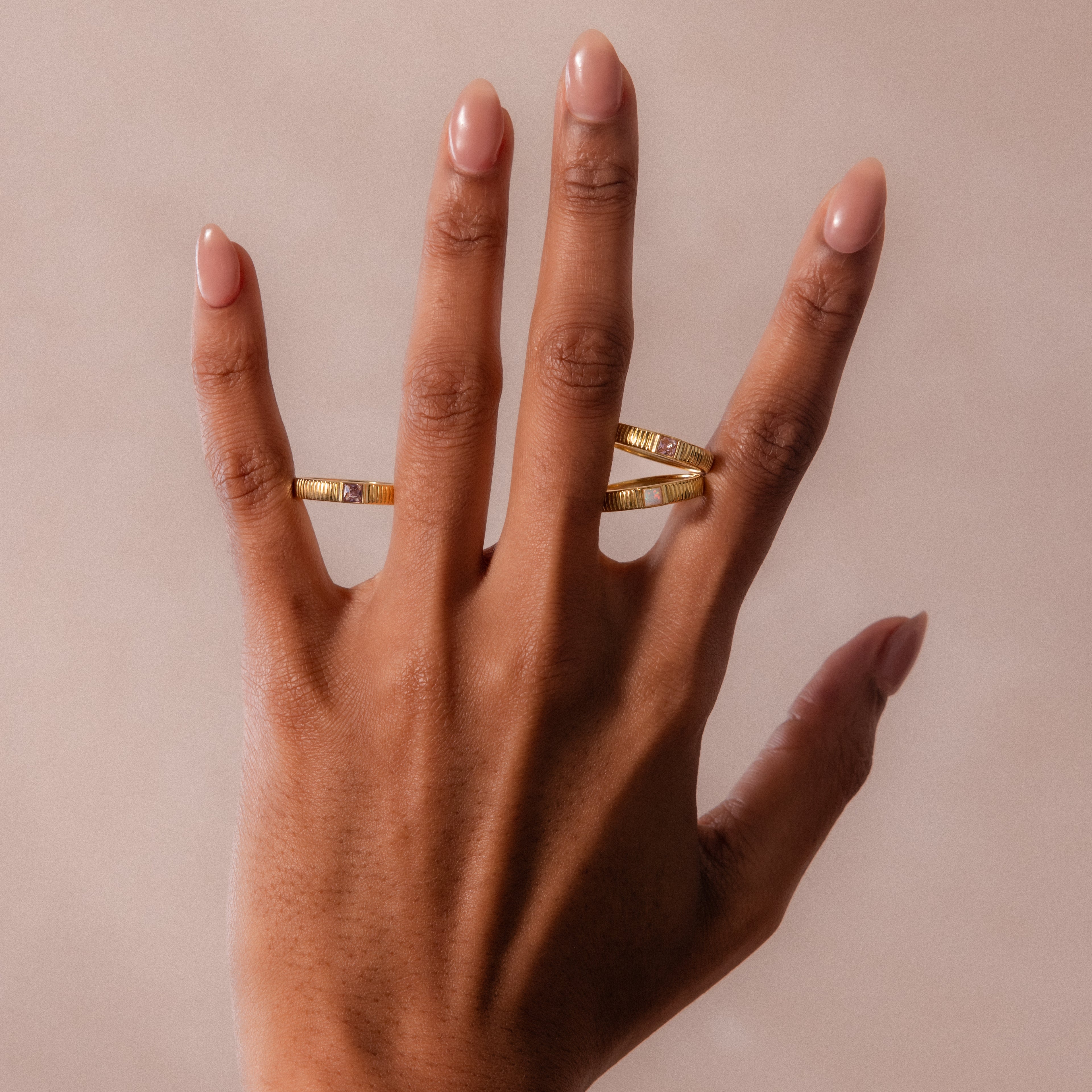 A hand with nude nails wears the Ribbed Peridot Band, a modern gold ring, on the middle finger and a square birthstone ring on the ring finger, set against a neutral background.