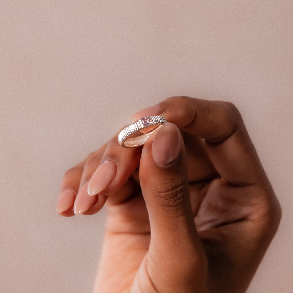A hand with manicured nails holds the Ribbed Alexandrite Band, a silver ring featuring a delicate square-cut alexandrite stone, against a neutral background.