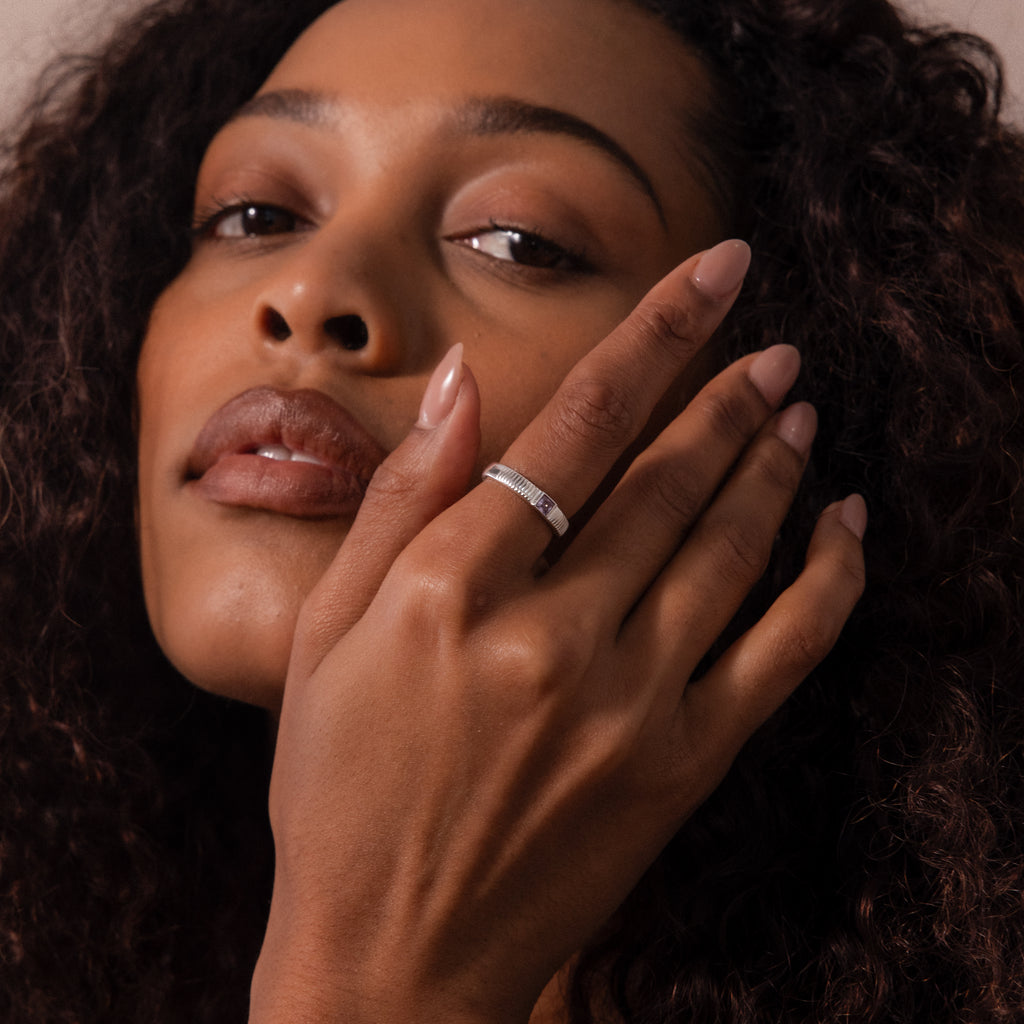 A woman with curly hair wears the Ribbed Alexandrite Band on her finger, touches her face, and looks at the camera.