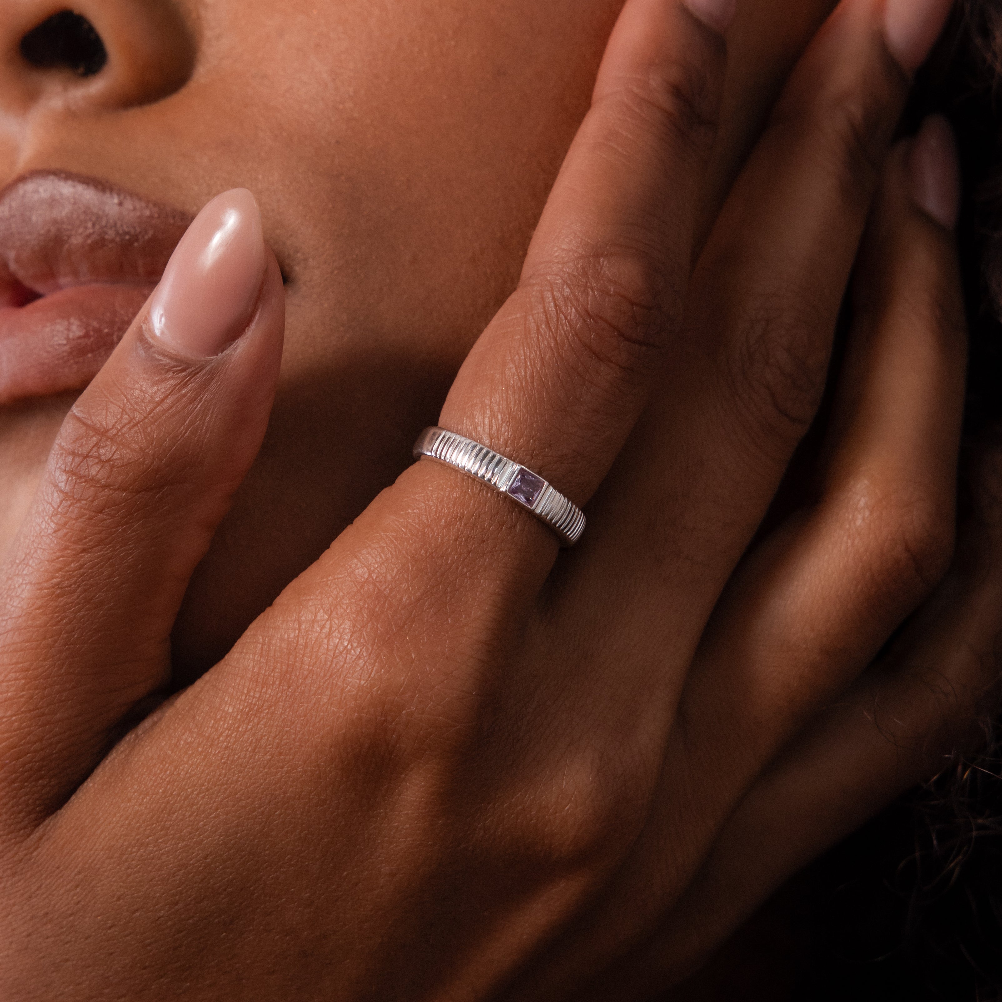 A close-up of a hand wearing the Ribbed Alexandrite Band gently touching the face of a person with smooth brown skin.
