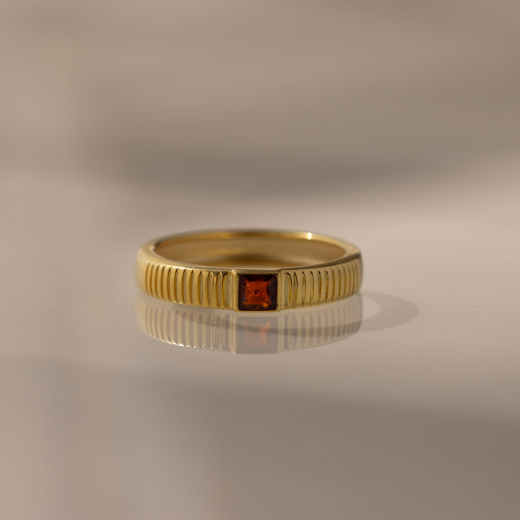 The Ribbed Garnet Band, a gold stacking ring with vertical grooves and a central square red gemstone, is displayed on a reflective surface.