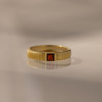 The Ribbed Garnet Band, a gold stacking ring with vertical grooves and a central square red gemstone, is displayed on a reflective surface.