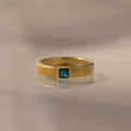 The Ribbed Blue Zircon Band, a gold stacking ring featuring a square blue gemstone at its center, is displayed on a reflective surface.