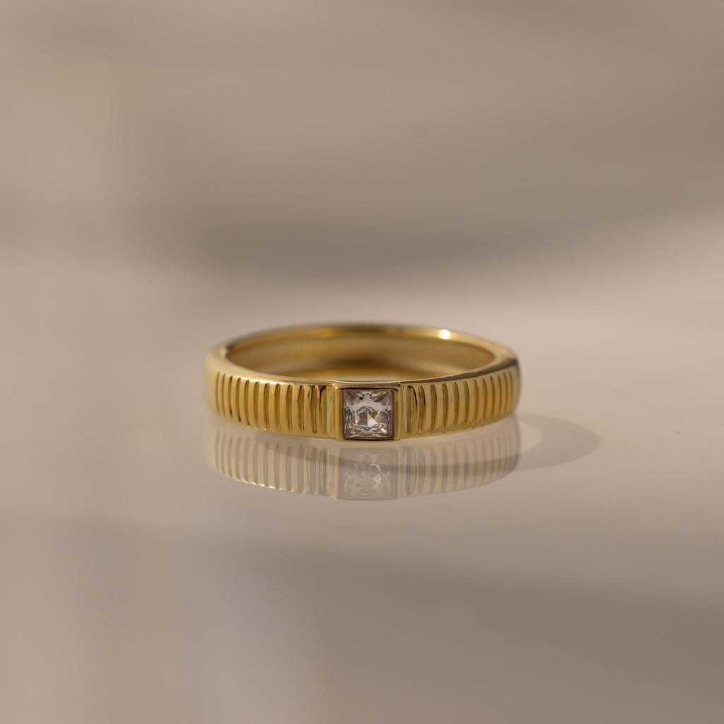 The Ribbed Diamond Band features gold ribbing and a square-cut center diamond, perfect for stacking or wearing alone. Shown on a neutral background.