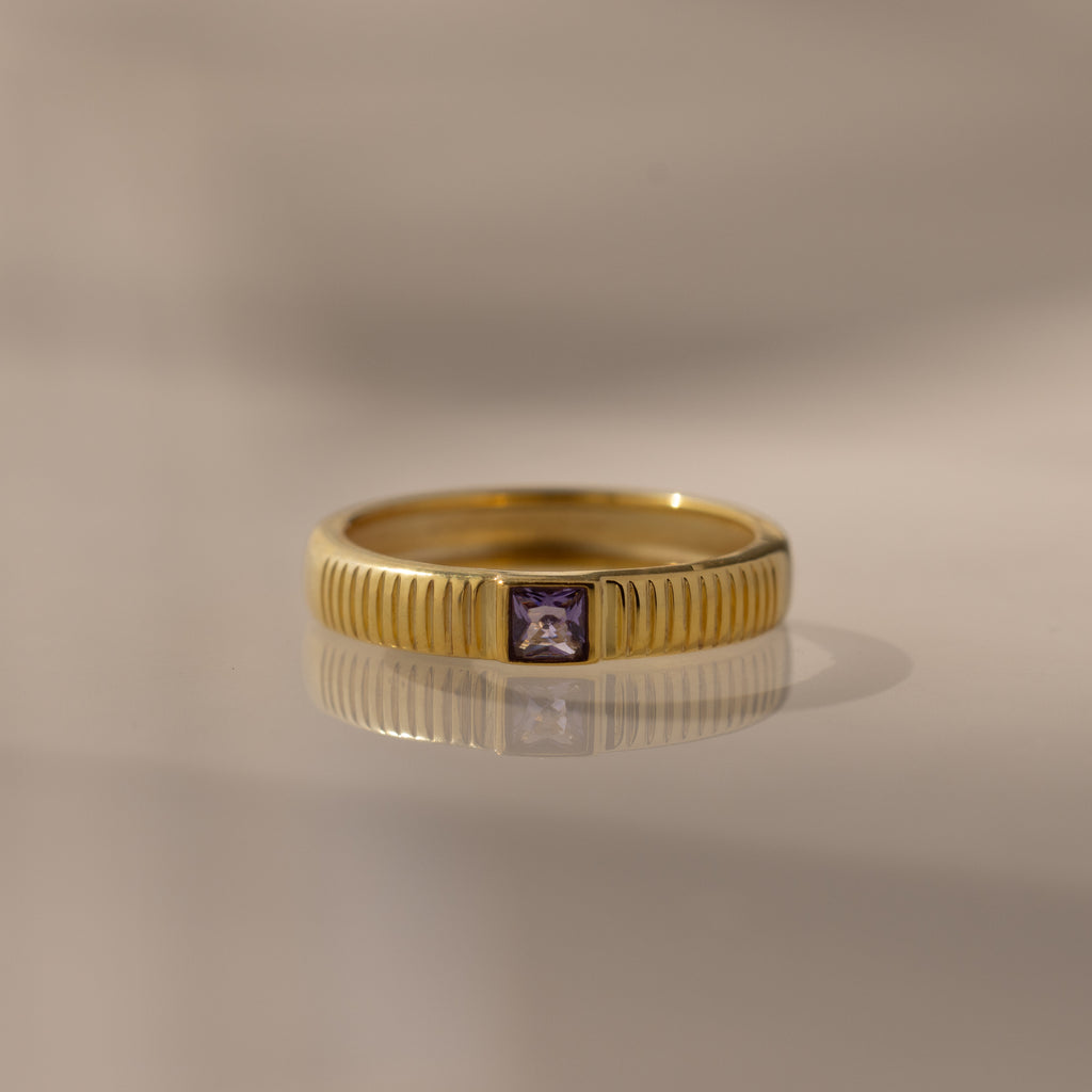 The Ribbed Amethyst Band is a gold stacking ring with vertical grooves and a small square purple amethyst in the center, shown on a reflective surface.