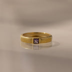 The Ribbed Amethyst Band is a gold stacking ring with vertical grooves and a small square purple amethyst in the center, shown on a reflective surface.