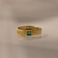 The Ribbed Emerald Band, featuring a gold ribbed design and a square-cut emerald, is displayed on a reflective surface with a soft background.