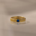 The Ribbed Sapphire Band features a gold ribbed, textured band with a small square blue sapphire at the center, ideal as a birthstone ring and shown on a neutral background.