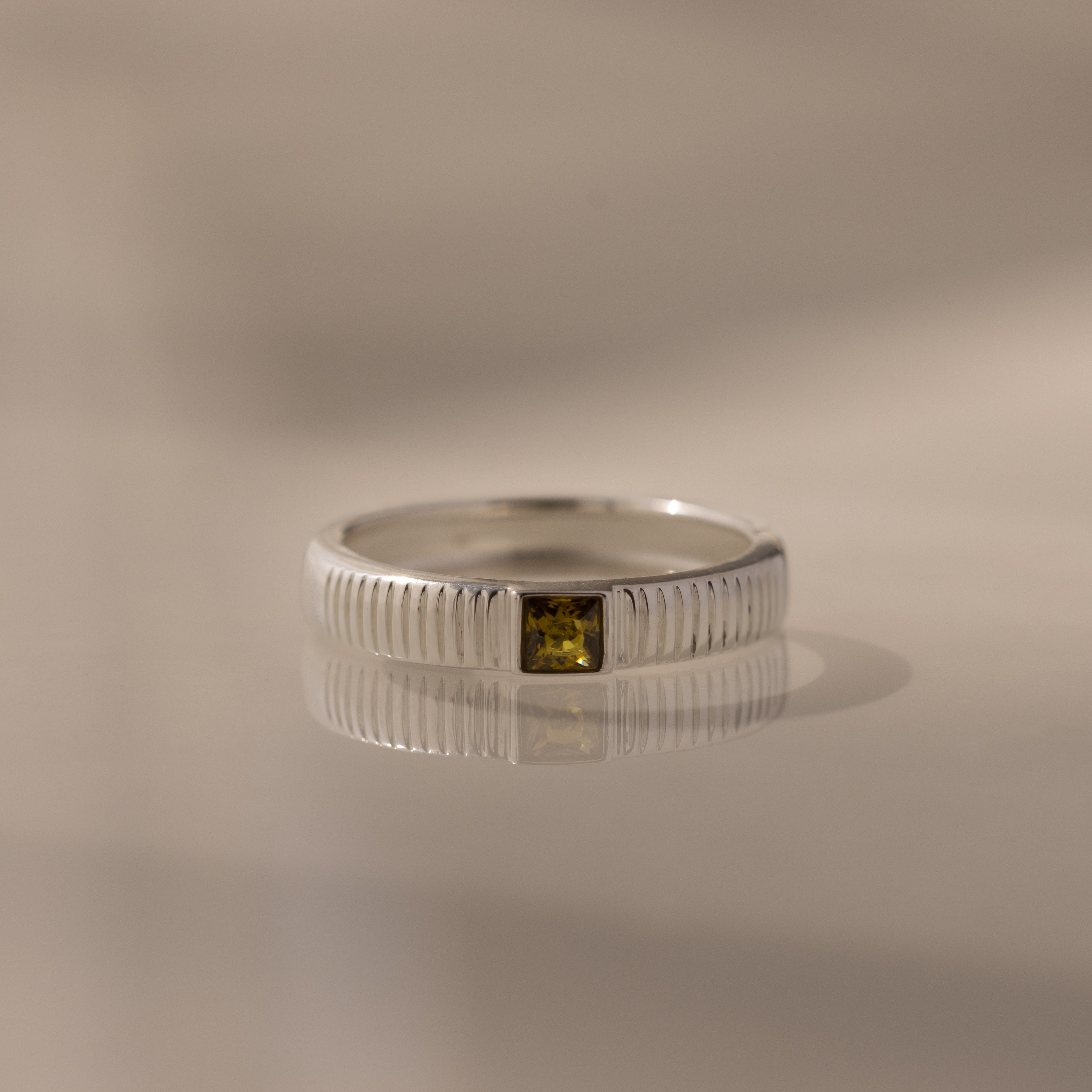 The Ribbed Peridot Band is a silver ring featuring a small, square-cut yellow-green peridot stone, shown on a reflective surface.