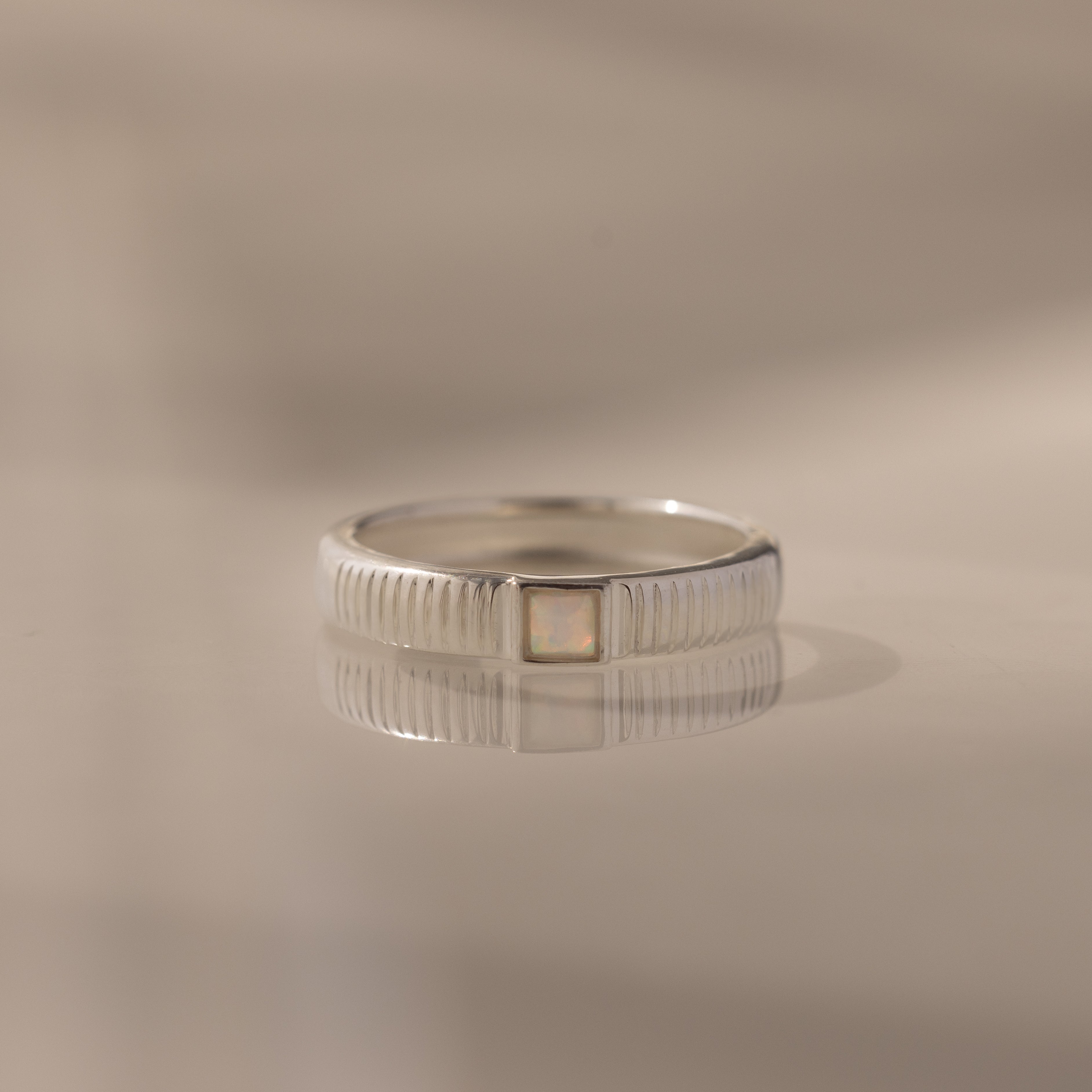 The Ribbed Opal Band is a silver ring with a square opal stone, engraved vertical lines, and a ribbed band, displayed on a reflective surface.