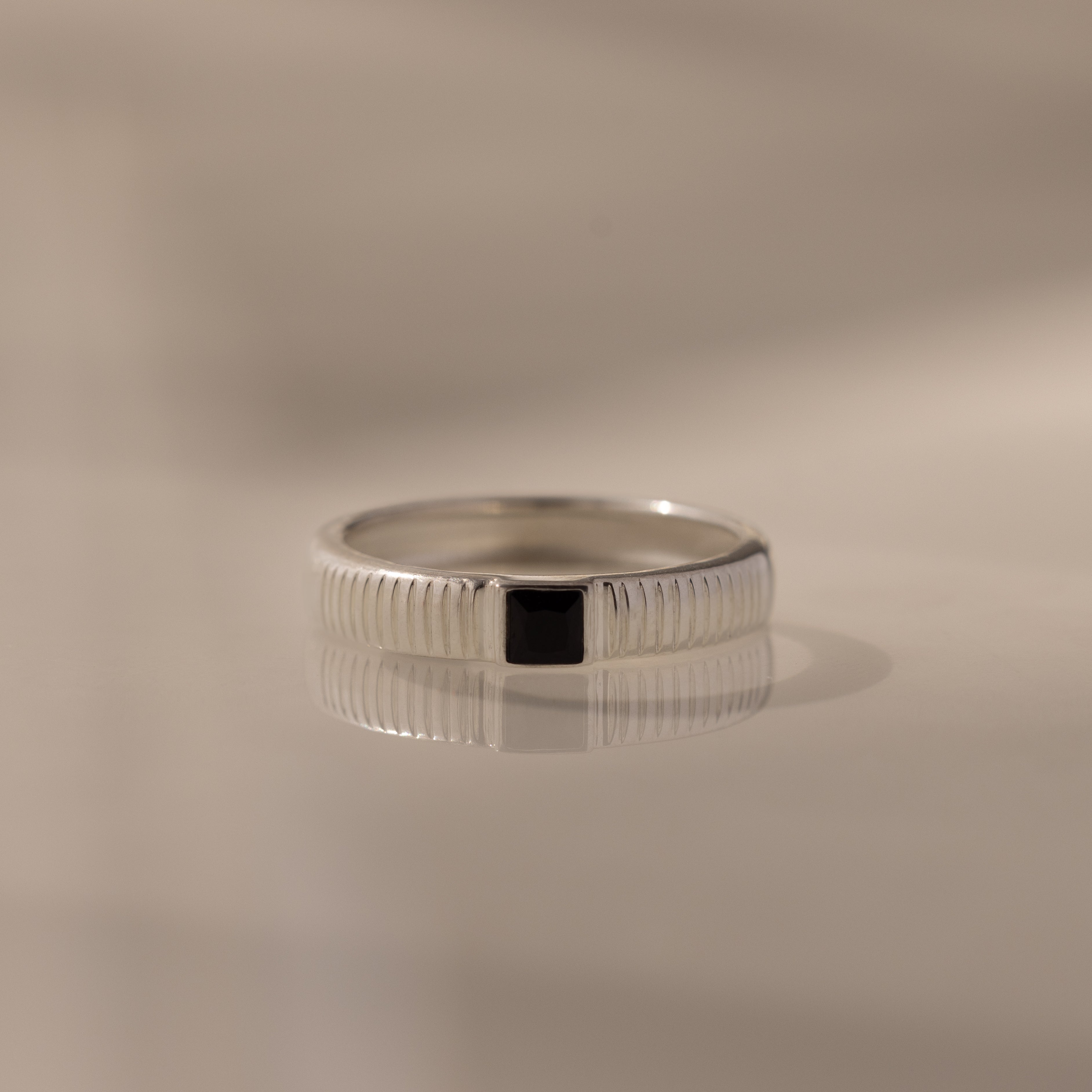 The Ribbed Onyx Band is a silver ring with a square-cut black onyx set at the center, shown on a reflective surface.
