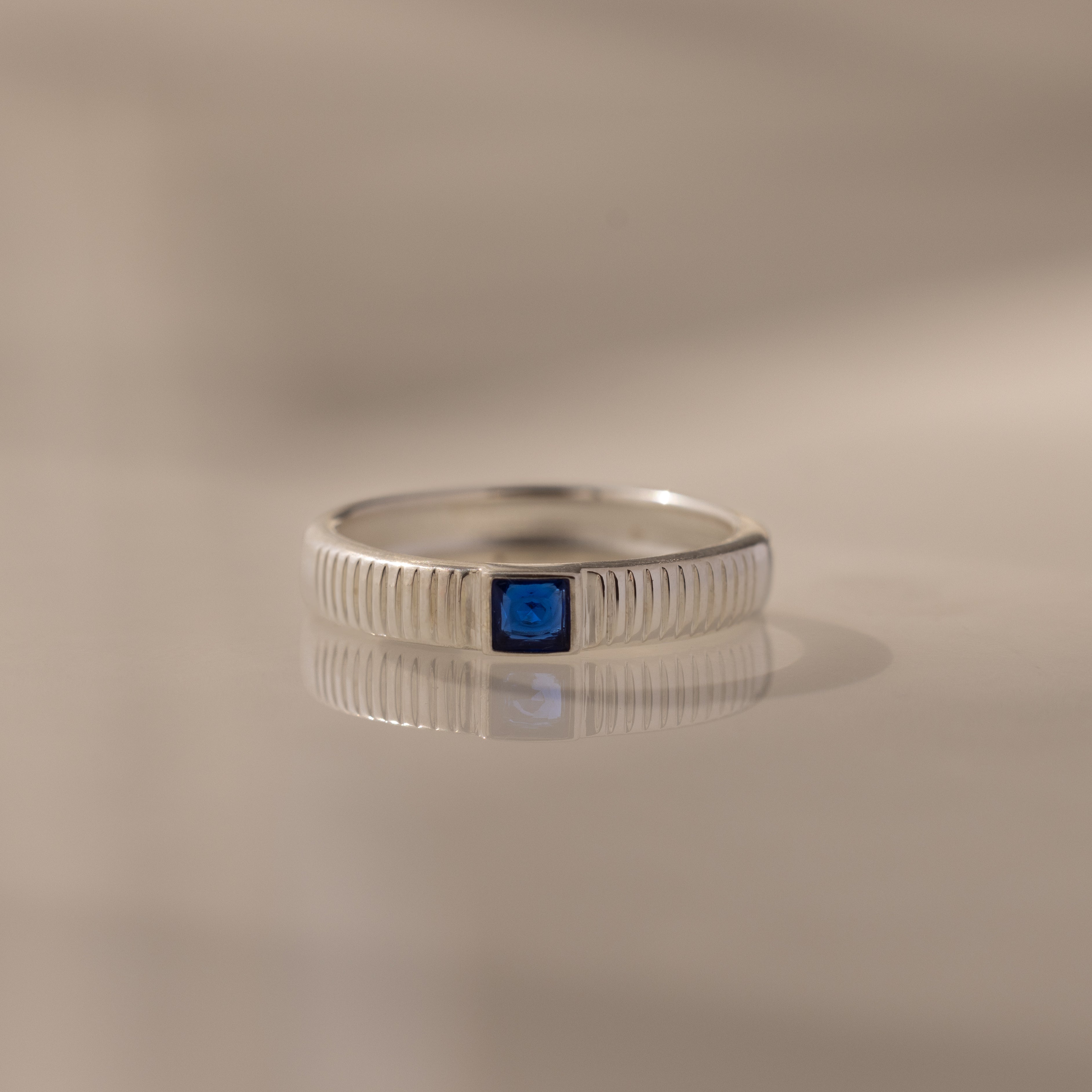 The Ribbed Sapphire Band is a silver stacking ring with a ridged band and a small blue square gemstone, shown on a smooth surface.
