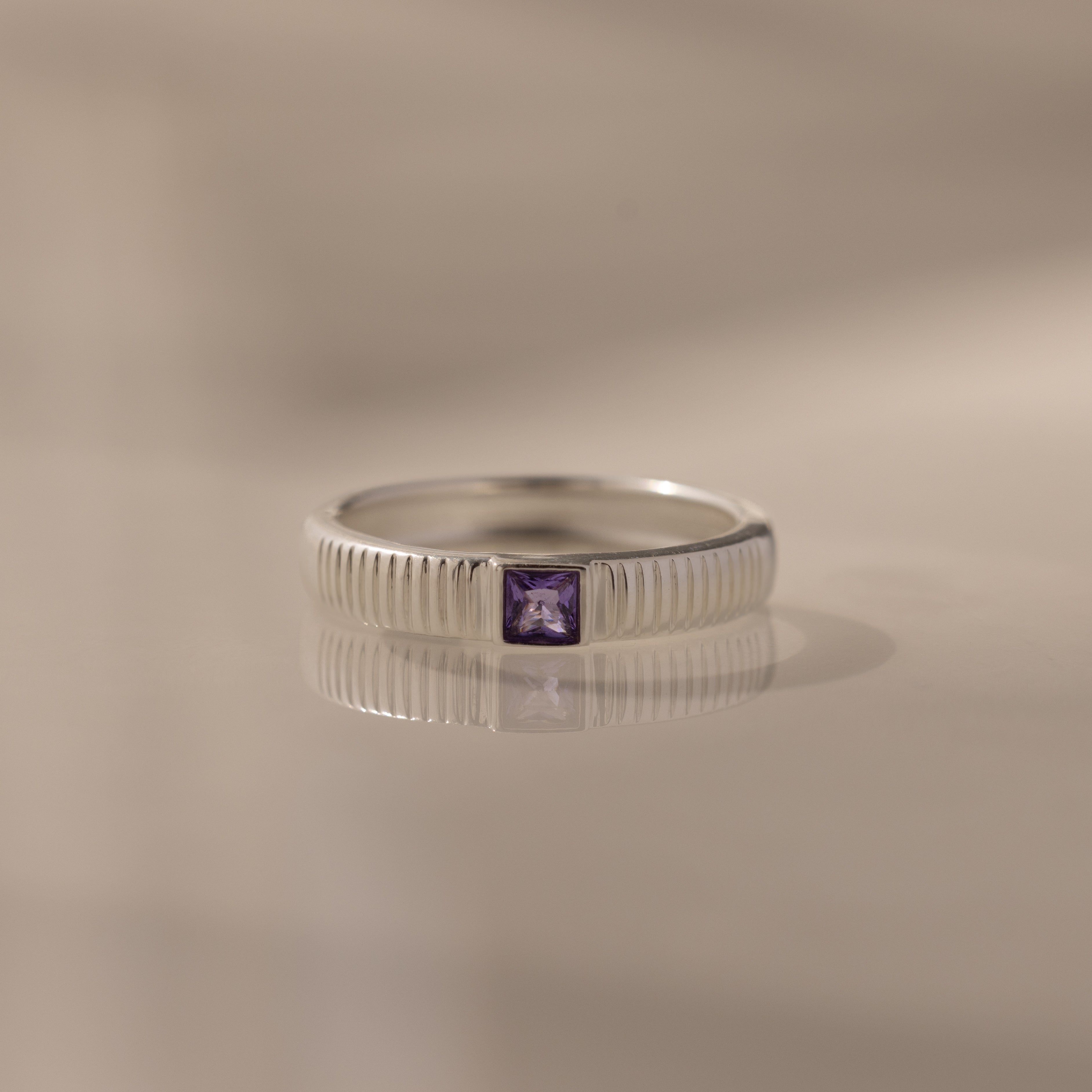 The Ribbed Amethyst Band features vertical grooves and a square purple gemstone at its center, displayed on a reflective surface.