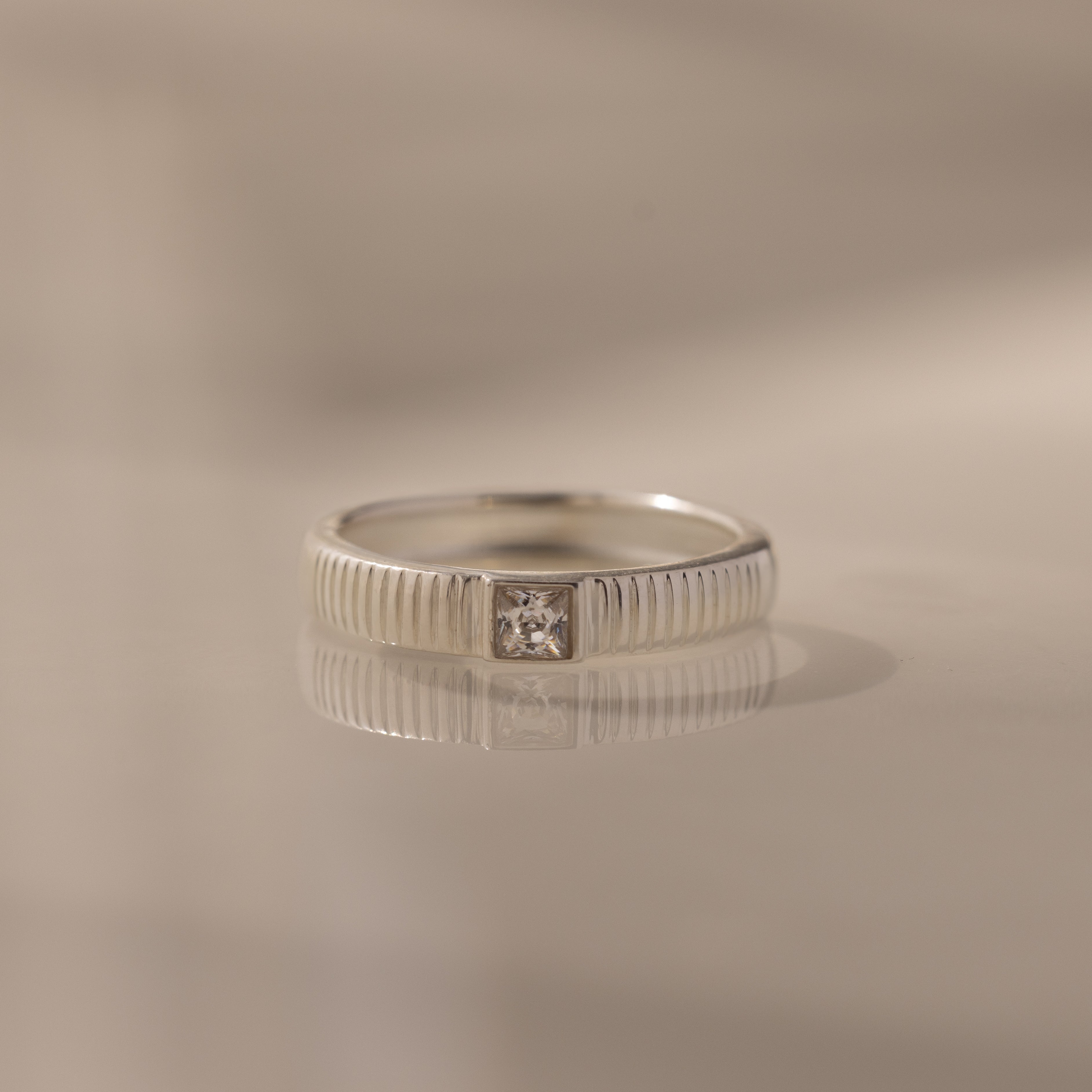 The Ribbed Diamond Band is a silver stacking ring featuring a grooved band and a square-cut diamond centerpiece, displayed on a reflective surface.