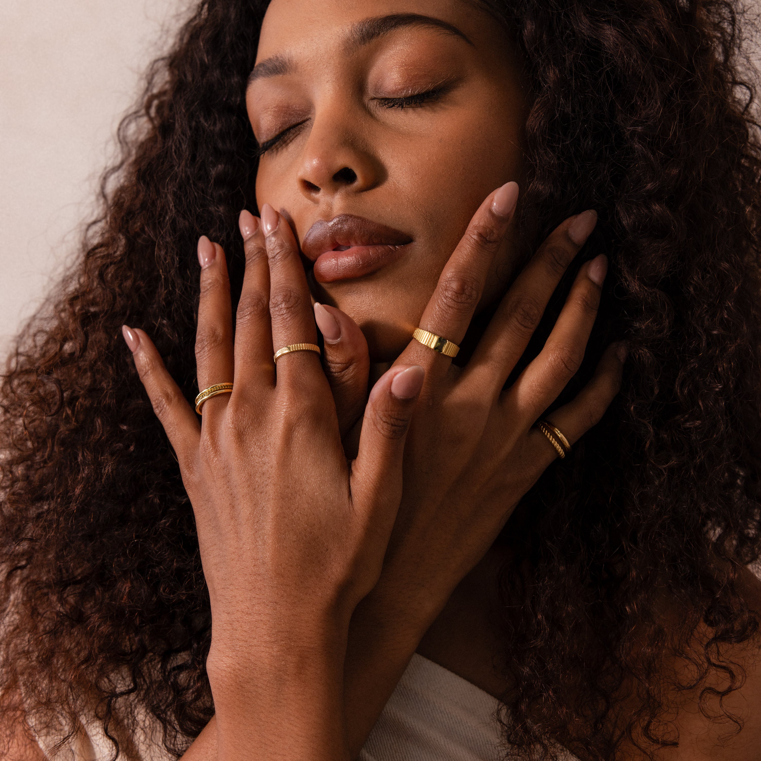 A woman with curly hair gently touches her face, showcasing the Ribbed Name Ring in 18K Gold and other gold bands—a refined display of personalized jewelry.