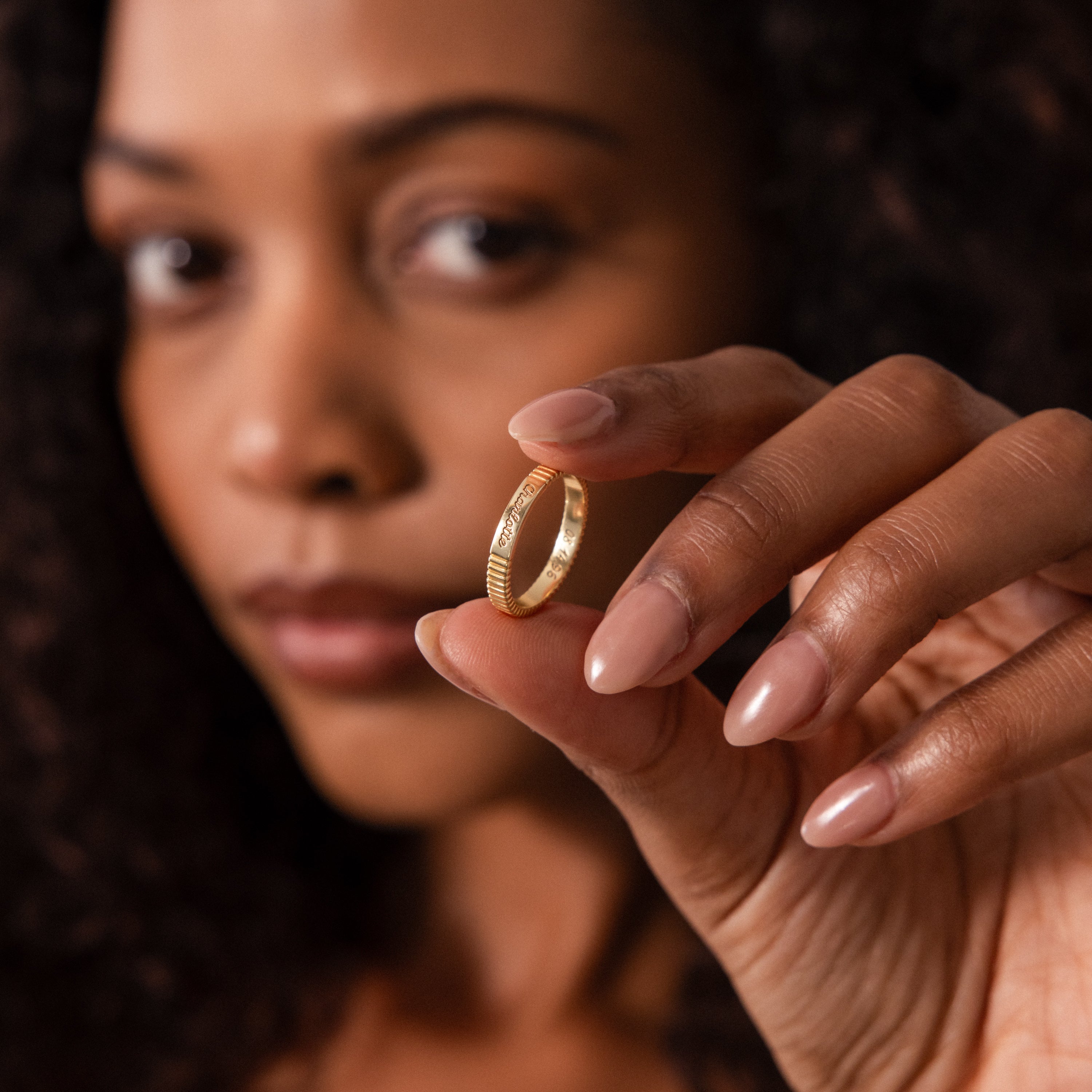 A woman with nude nails holds the Ribbed Name Ring in 18K Gold between her fingers, her face softly blurred in the background.