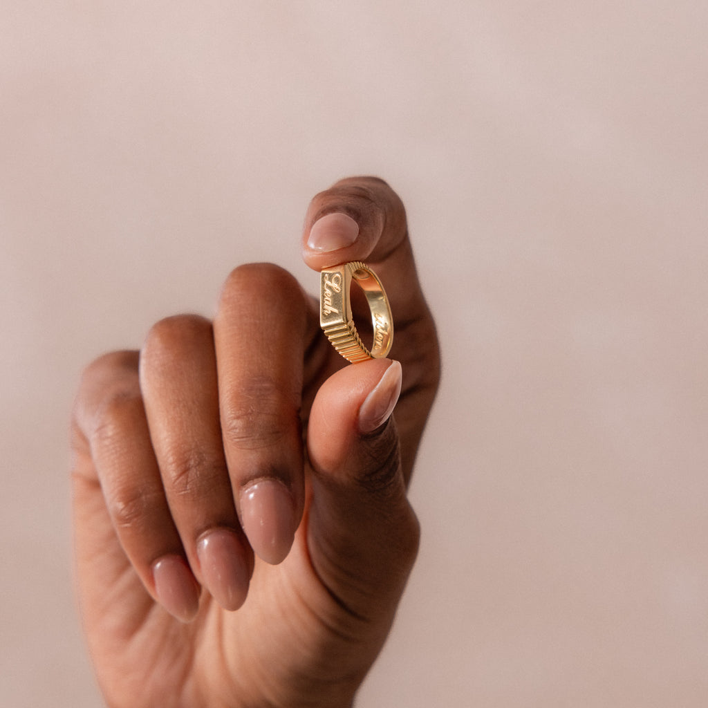 A hand with neutral nail polish holds the Ribbed Name Signet Ring in 18K Gold, its bold design standing out against a soft beige background.