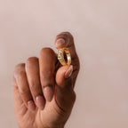 A hand with neutral nail polish holds the Ribbed Name Signet Ring in 18K Gold, its bold design standing out against a soft beige background.