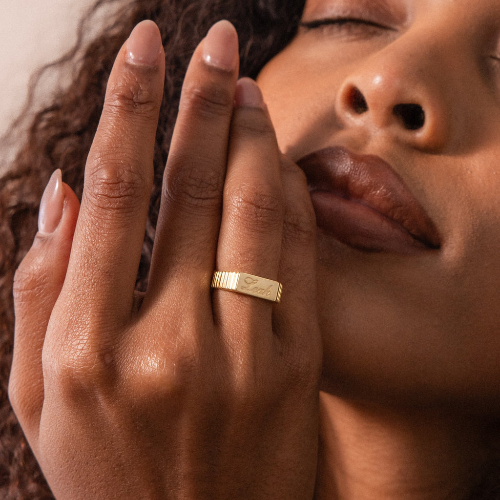 With her eyes closed, a woman raises her hand near her face to display the Ribbed Name Signet Ring—a gold statement ring engraved with "Faith.