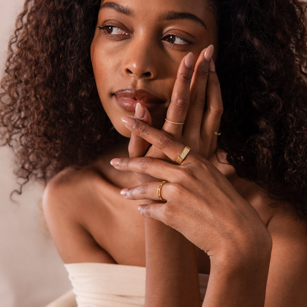 A woman with curly hair poses with her hands near her face, thoughtfully looking to the side and showcasing the bold Ribbed Name Signet Ring in 18K Gold.