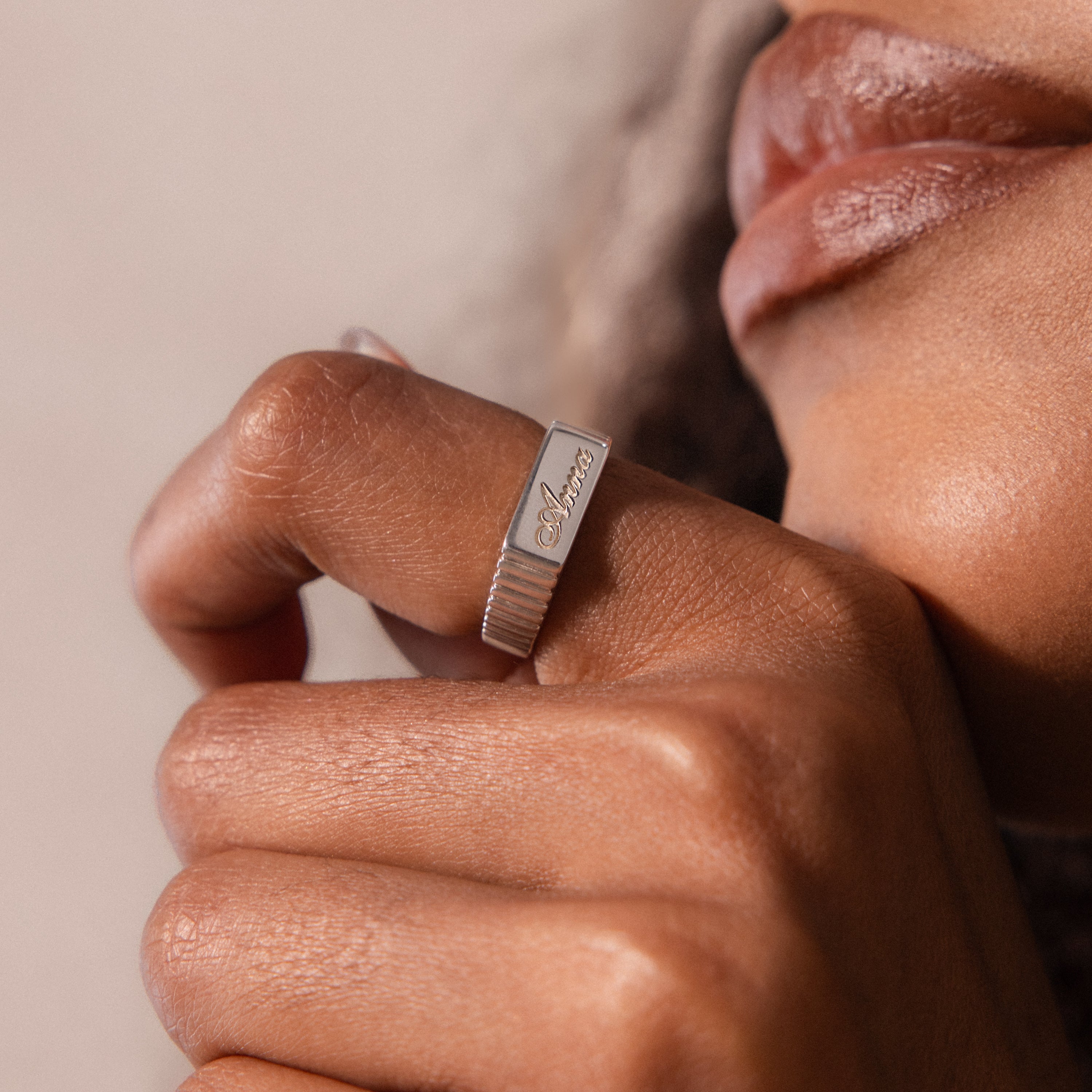 A close-up shows a hand near a person's lips wearing the Ribbed Name Signet Ring, engraved with text.