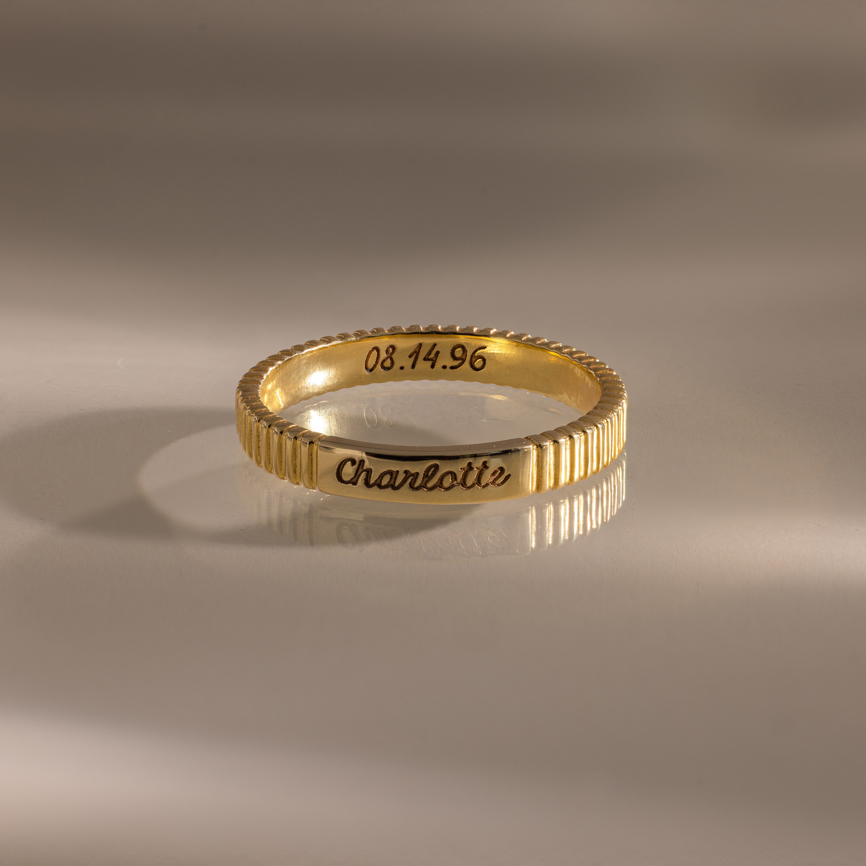 A Ribbed Name Ring engraved with "Charlotte" and the date 08.14.96 rests on a reflective surface, highlighting the unique personalization of this ring.