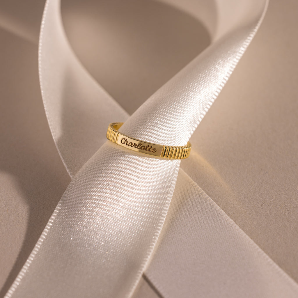 The Ribbed Name Ring in 18K Gold, engraved with 'Charlotte', rests on a white satin ribbon, highlighting the elegance of personalized jewelry.