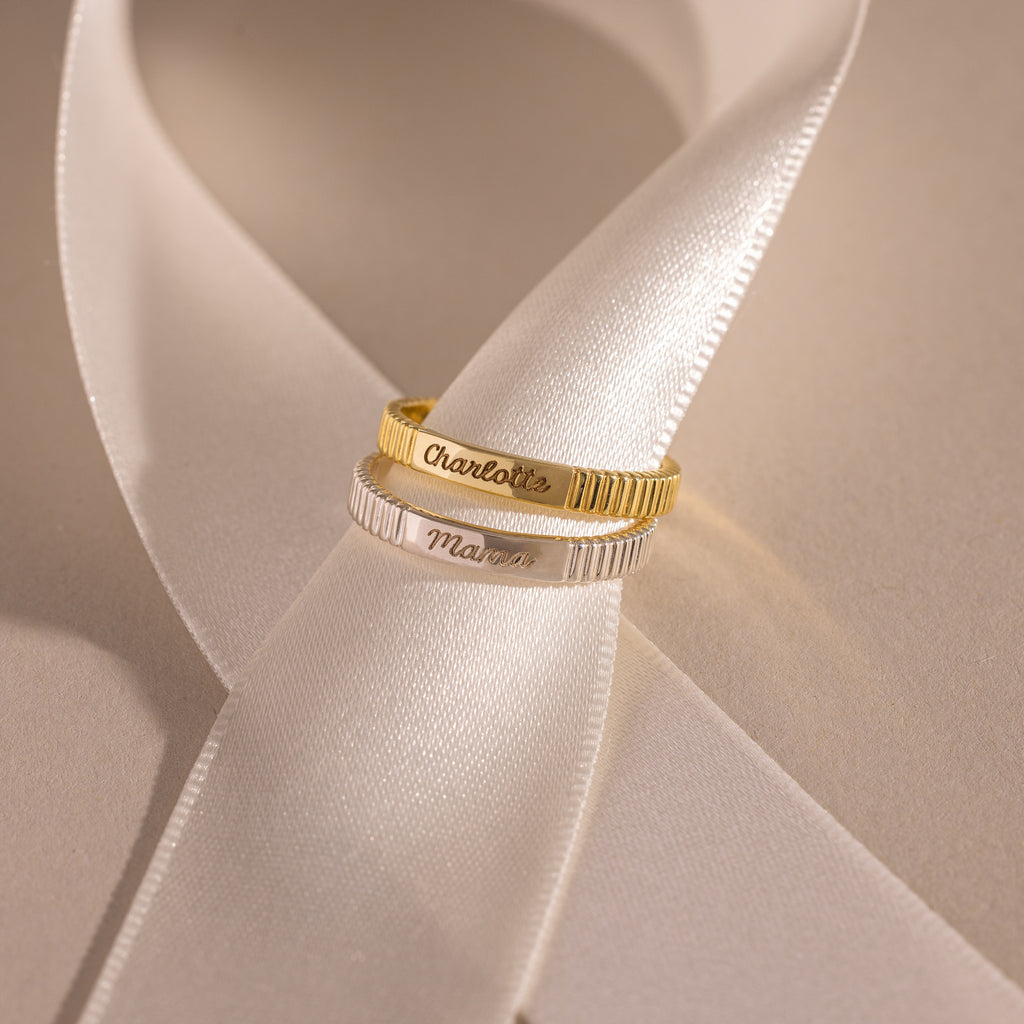 The Ribbed Name Ring, available in gold or silver, shines on a white satin ribbon under soft light—ideal as a personalized gift or custom engraved keepsake.