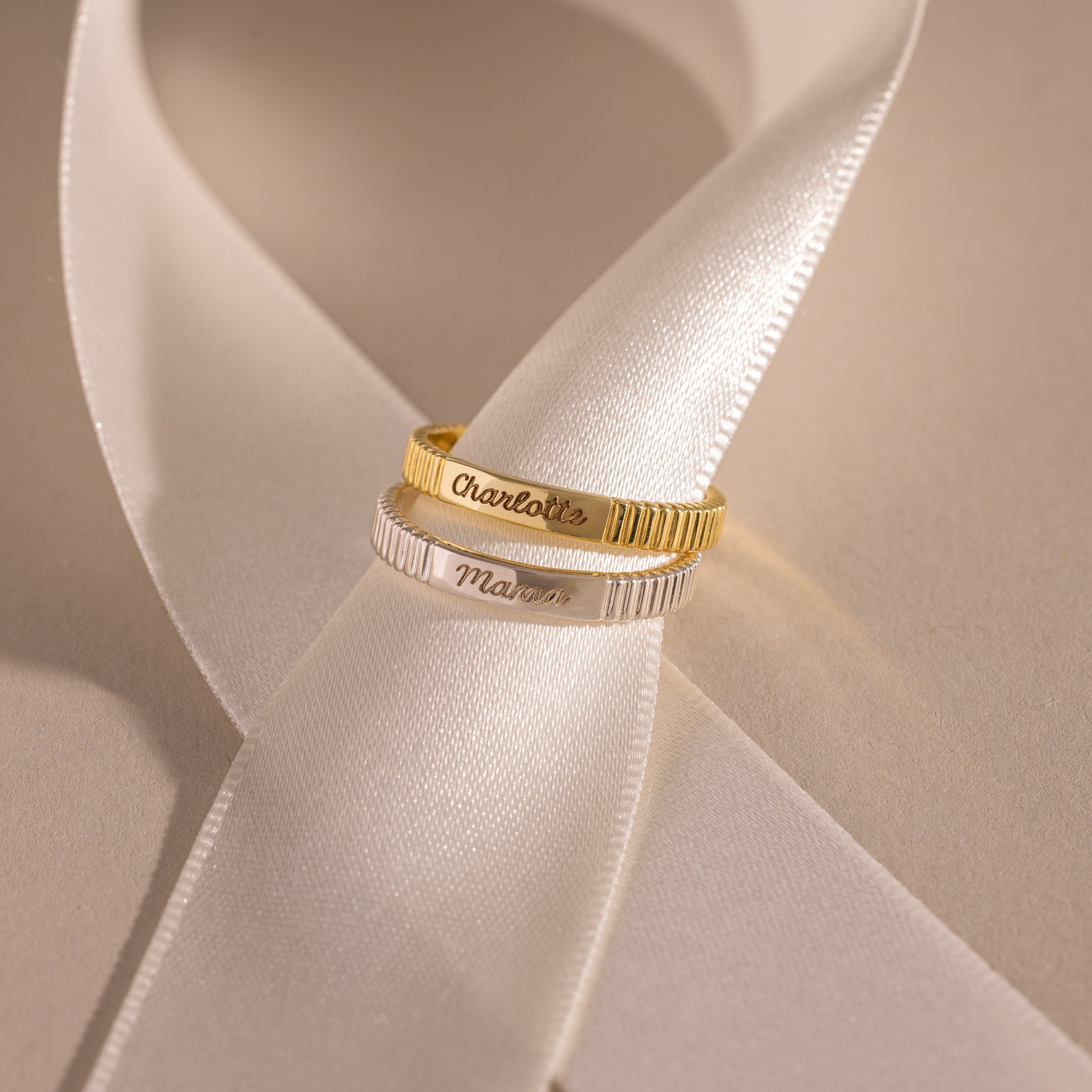 The Ribbed Name Ring, available in gold or silver, shines on a white satin ribbon under soft light—ideal as a personalized gift or custom engraved keepsake.