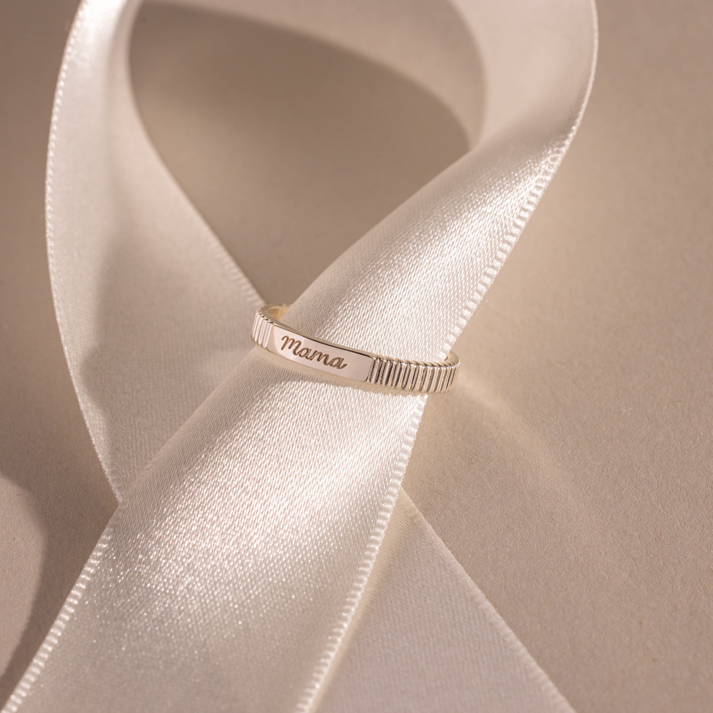 A silver Ribbed Name Ring engraved with Mama rests on a white satin ribbon.