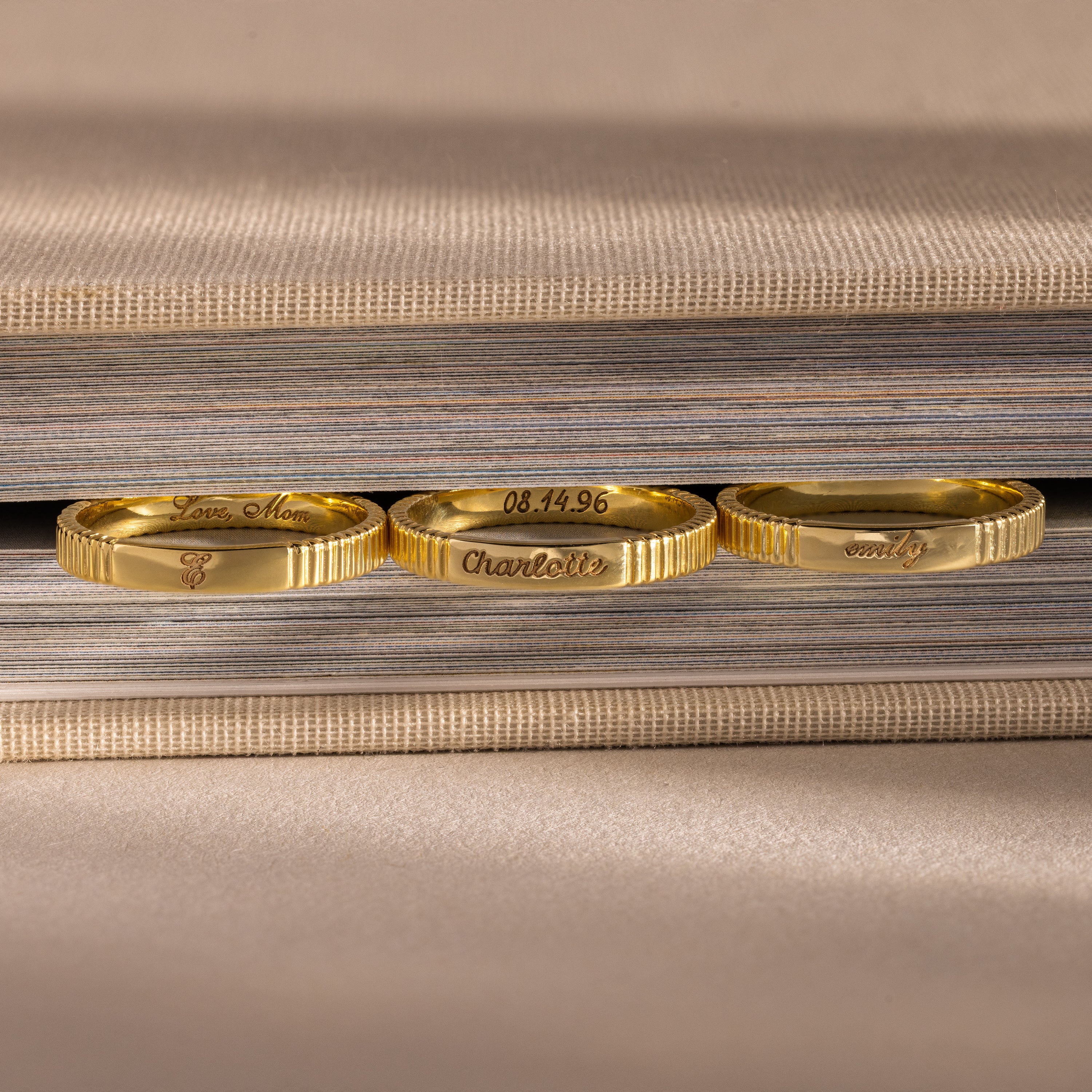 Three gold rings with engraved messages—including a custom piece and the Ribbed Name Ring in 18K Gold—rest upright against the edge of a closed book.