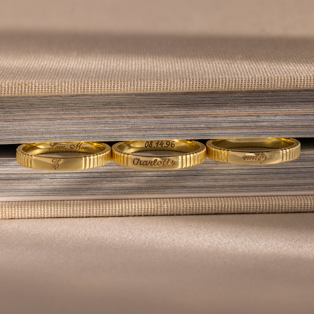 Three Ribbed Name Rings, each engraved with a custom name, rest on the edge of a closed, textured book.