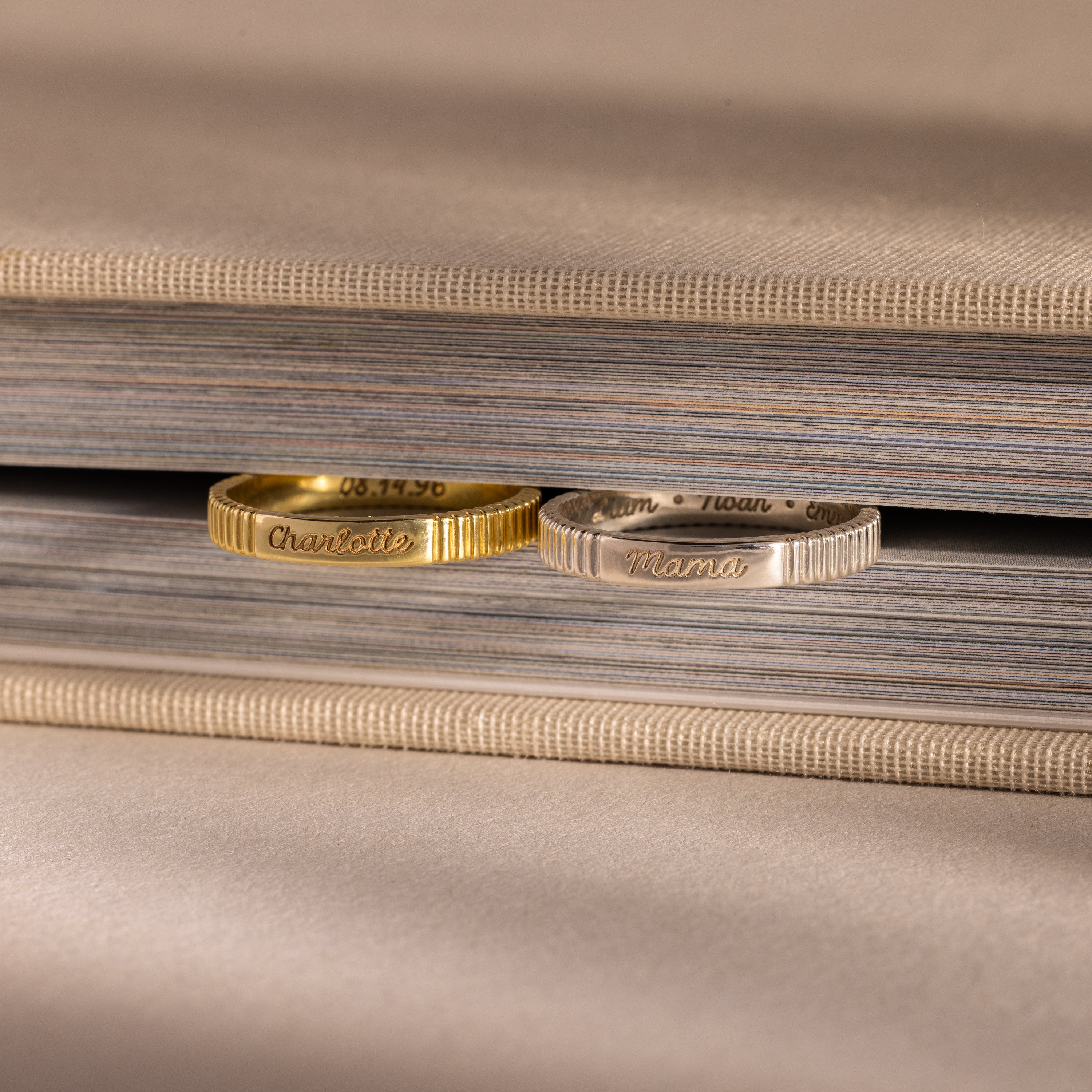 A gold Ribbed Name Ring and a silver engraved ring rest side by side on the edge of a closed beige book.