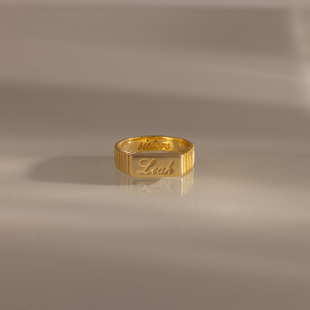 The Ribbed Name Signet Ring in 18K Gold showcases "Leah" and "victory" elegantly engraved on the signet face, set on a smooth, neutral surface—a true statement piece.