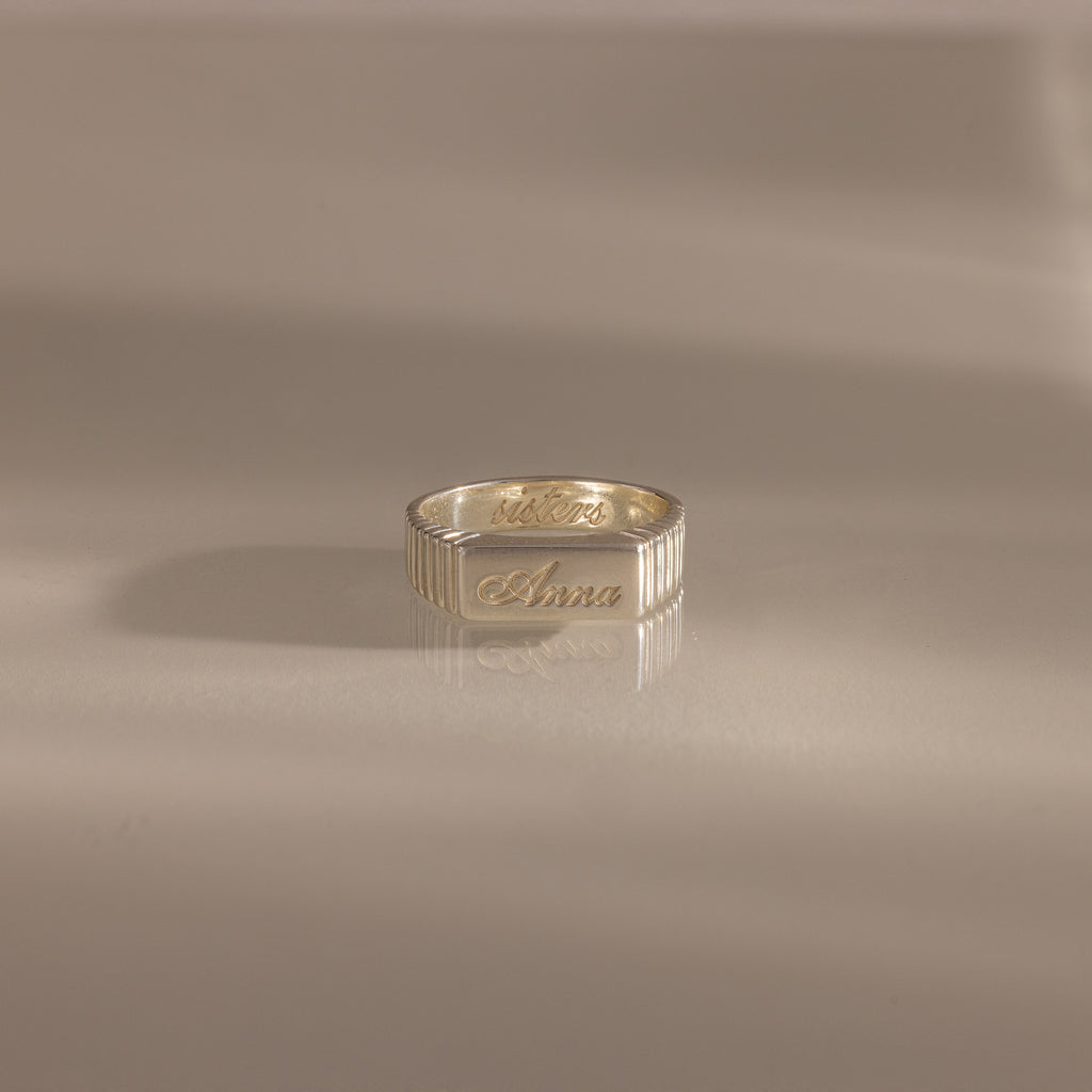 A Ribbed Name Signet Ring in Silver, engraved with the name Anna, rests on a smooth, light surface.