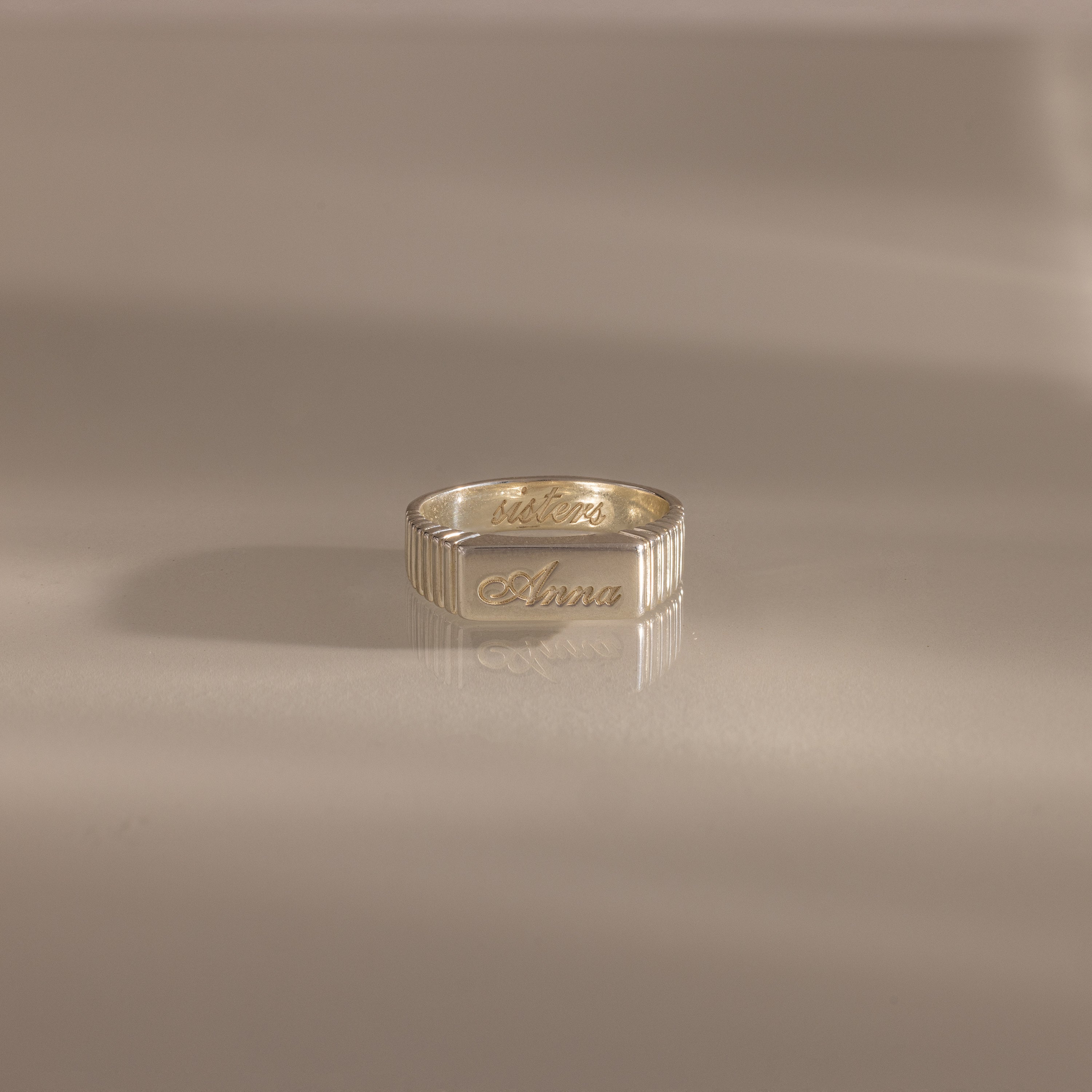 A Ribbed Name Signet Ring in Silver, engraved with the name Anna, rests on a smooth, light surface.