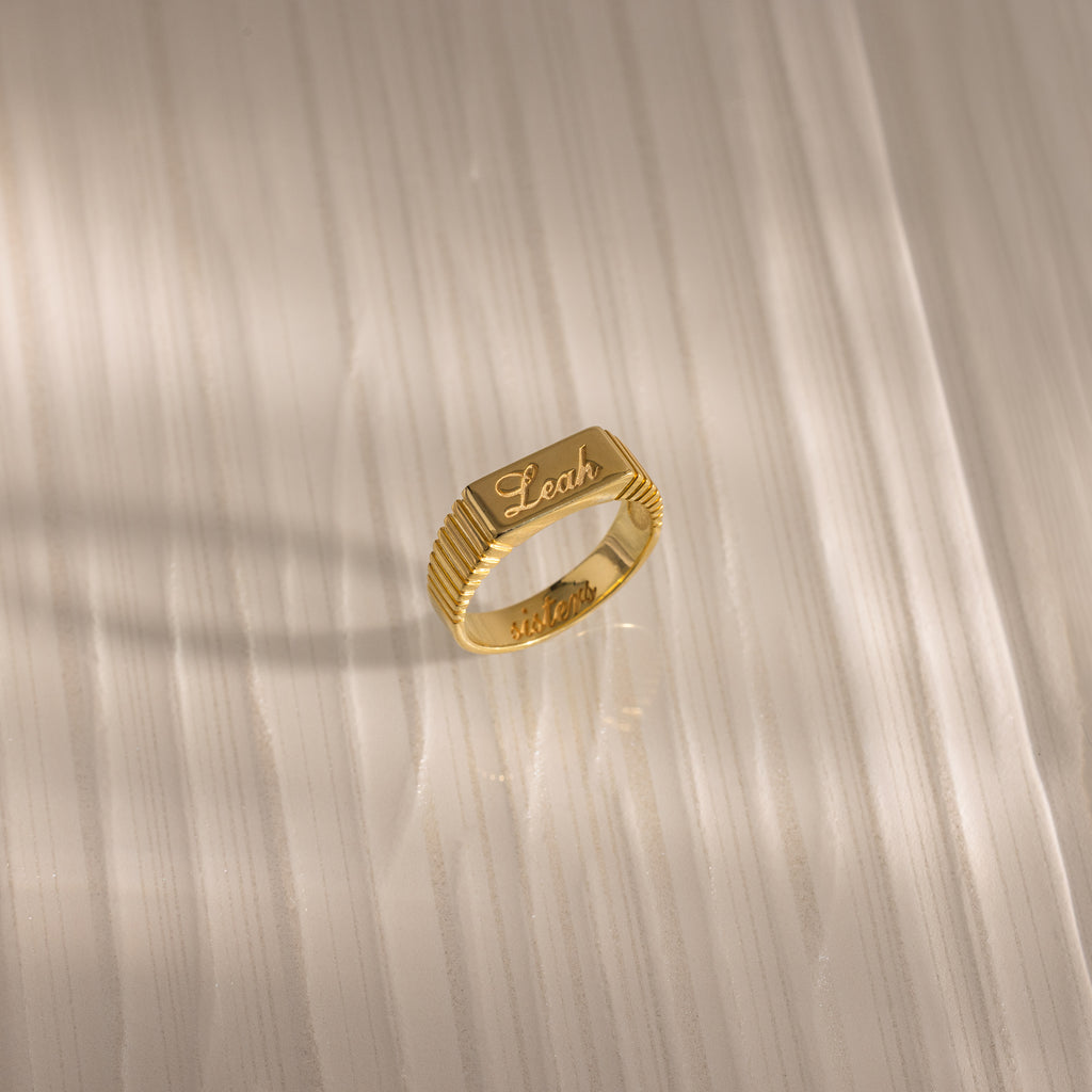 A Ribbed Name Signet Ring in 18K Gold engraved with the name Leah rests on a light-colored, striped surface in soft light.