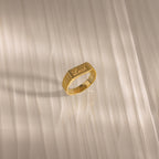 A Ribbed Name Signet Ring in 18K Gold engraved with the name Leah rests on a light-colored, striped surface in soft light.
