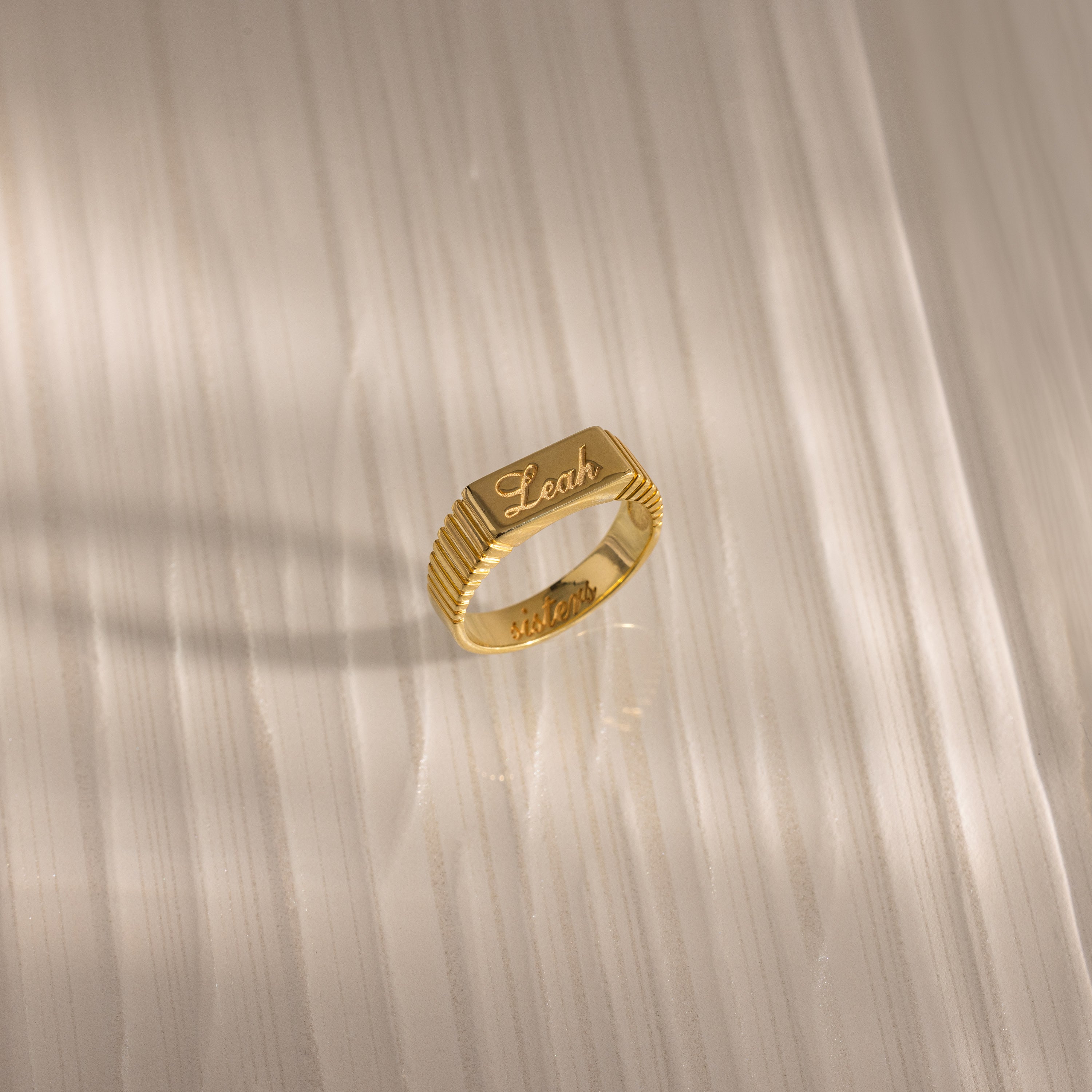 A Ribbed Name Signet Ring in 18K Gold engraved with the name Leah rests on a light-colored, striped surface in soft light.