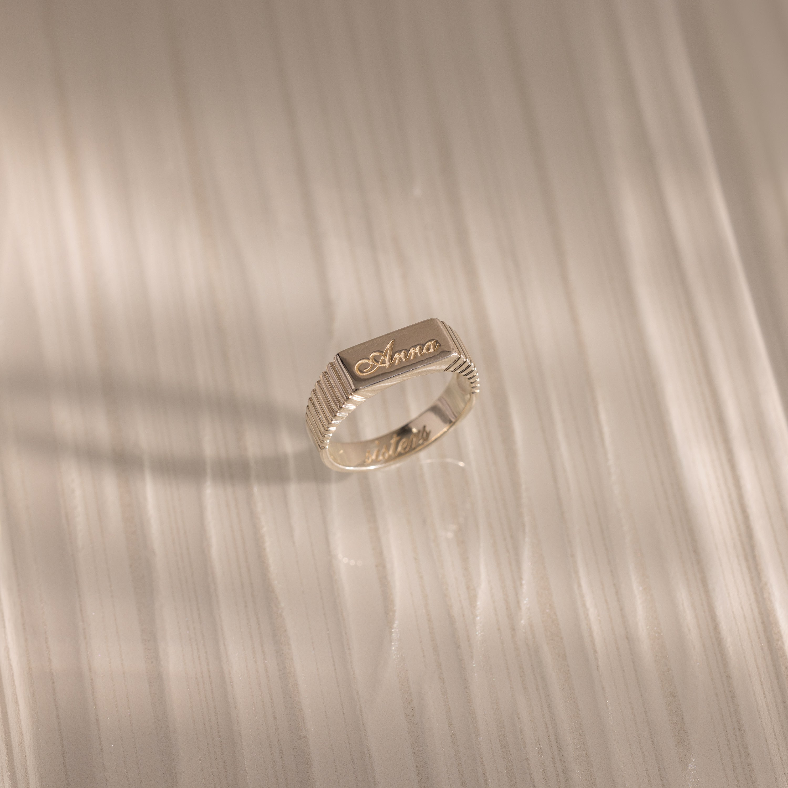 The Ribbed Name Signet Ring in Silver, engraved with the name Anna, rests on a light, textured wooden surface with soft shadows.