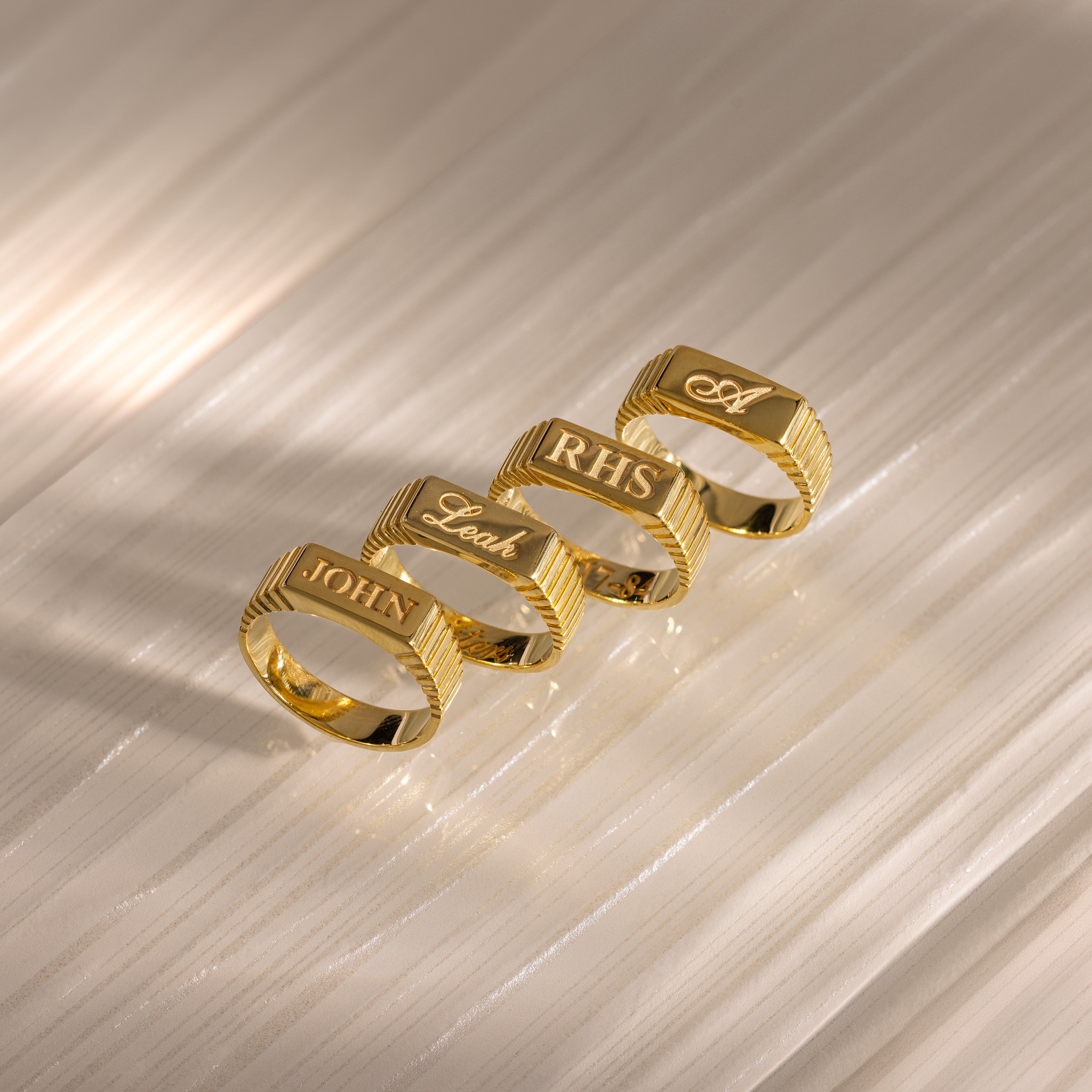 Four Ribbed Name Signet Rings in 18K Gold engraved with John, Leah, RHS, and A are lined up on a textured surface—each ring offering a unique blend of classic elegance and personal style.