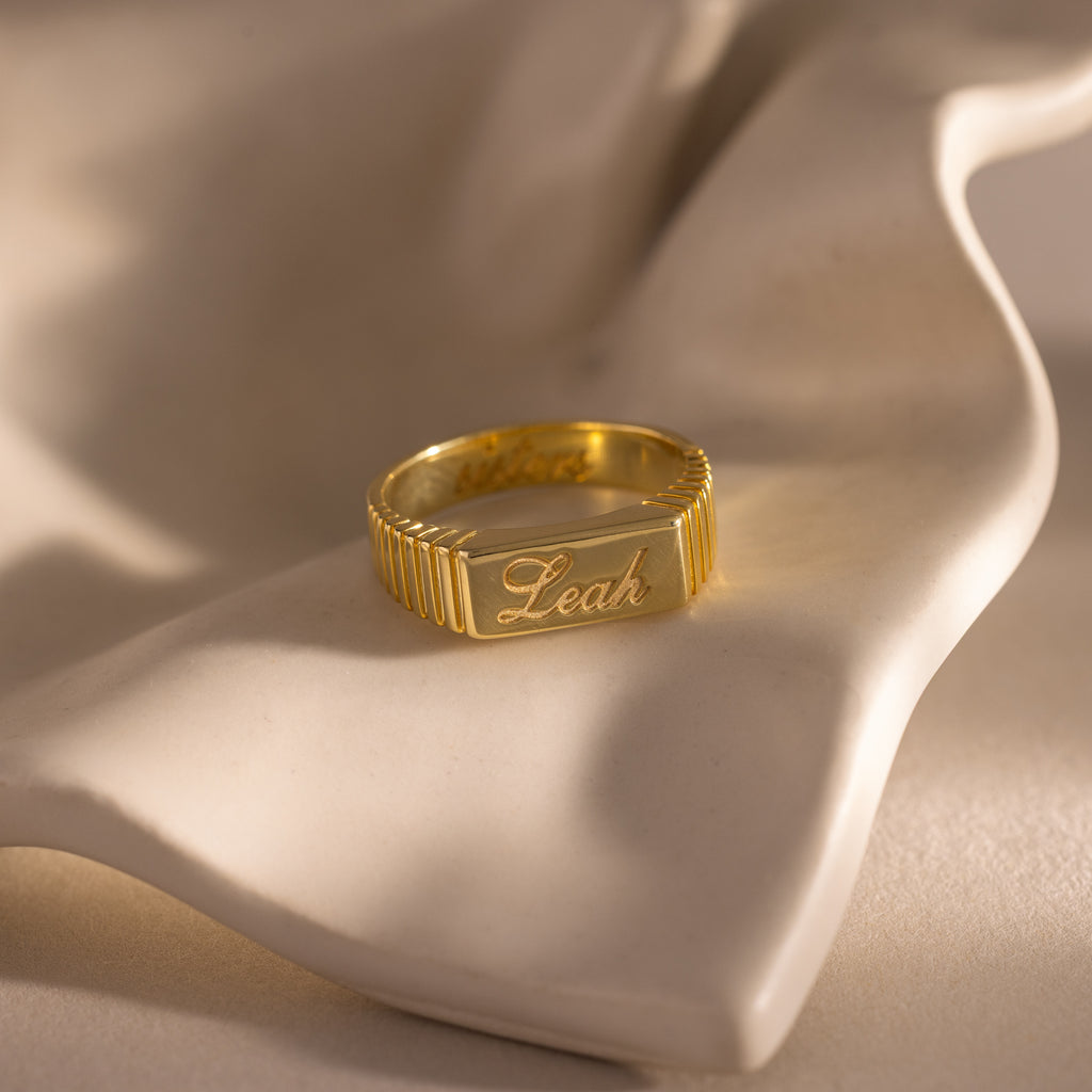 The Ribbed Name Signet Ring in 18K Gold, engraved with "Leah," rests on a soft white draped surface—a timeless statement ring with a beautifully detailed signet face.