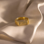 The Ribbed Name Signet Ring in 18K Gold, engraved with "Leah," rests on a soft white draped surface—a timeless statement ring with a beautifully detailed signet face.