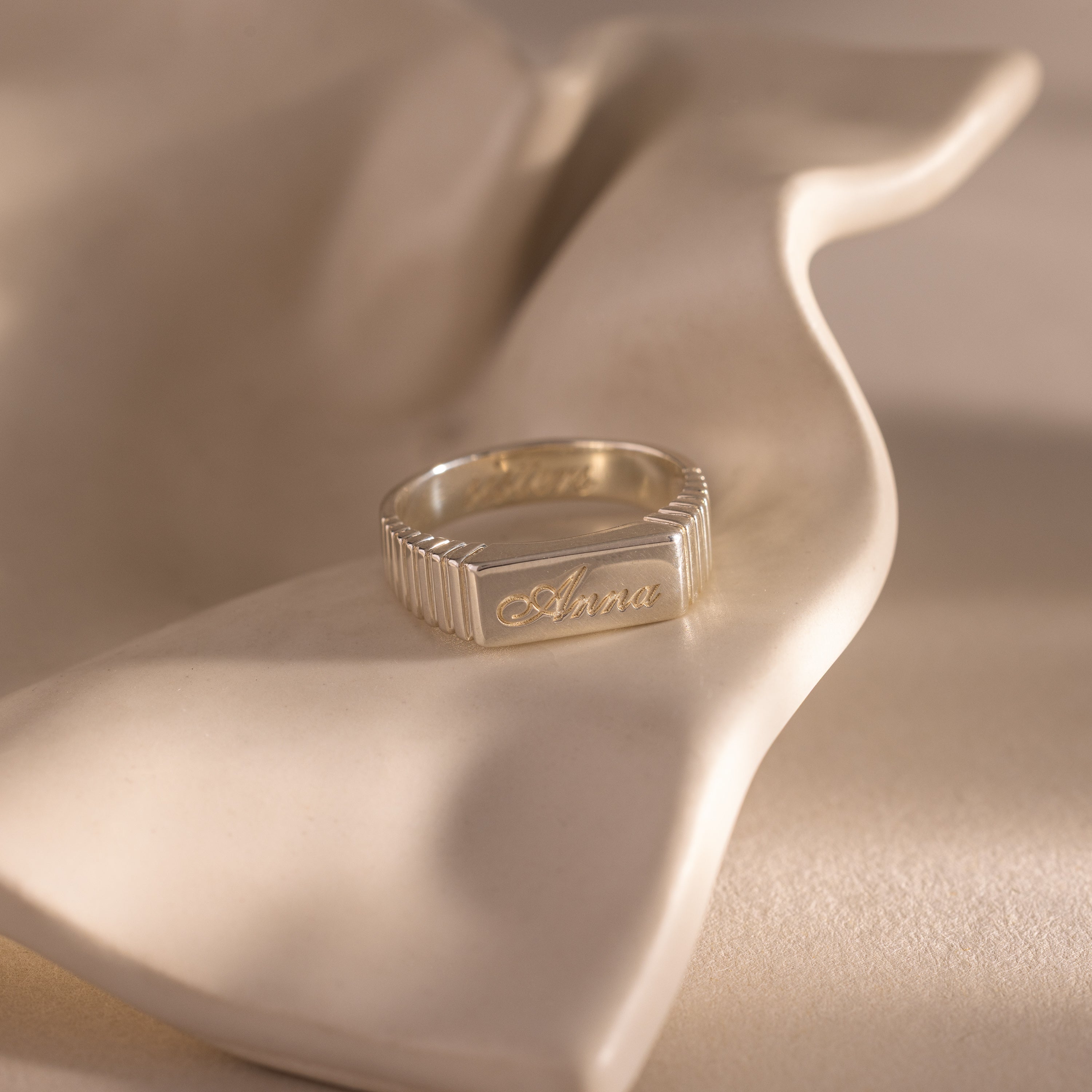 The Ribbed Name Signet Ring in Silver, engraved with the name Anna, rests on a softly curved beige surface.