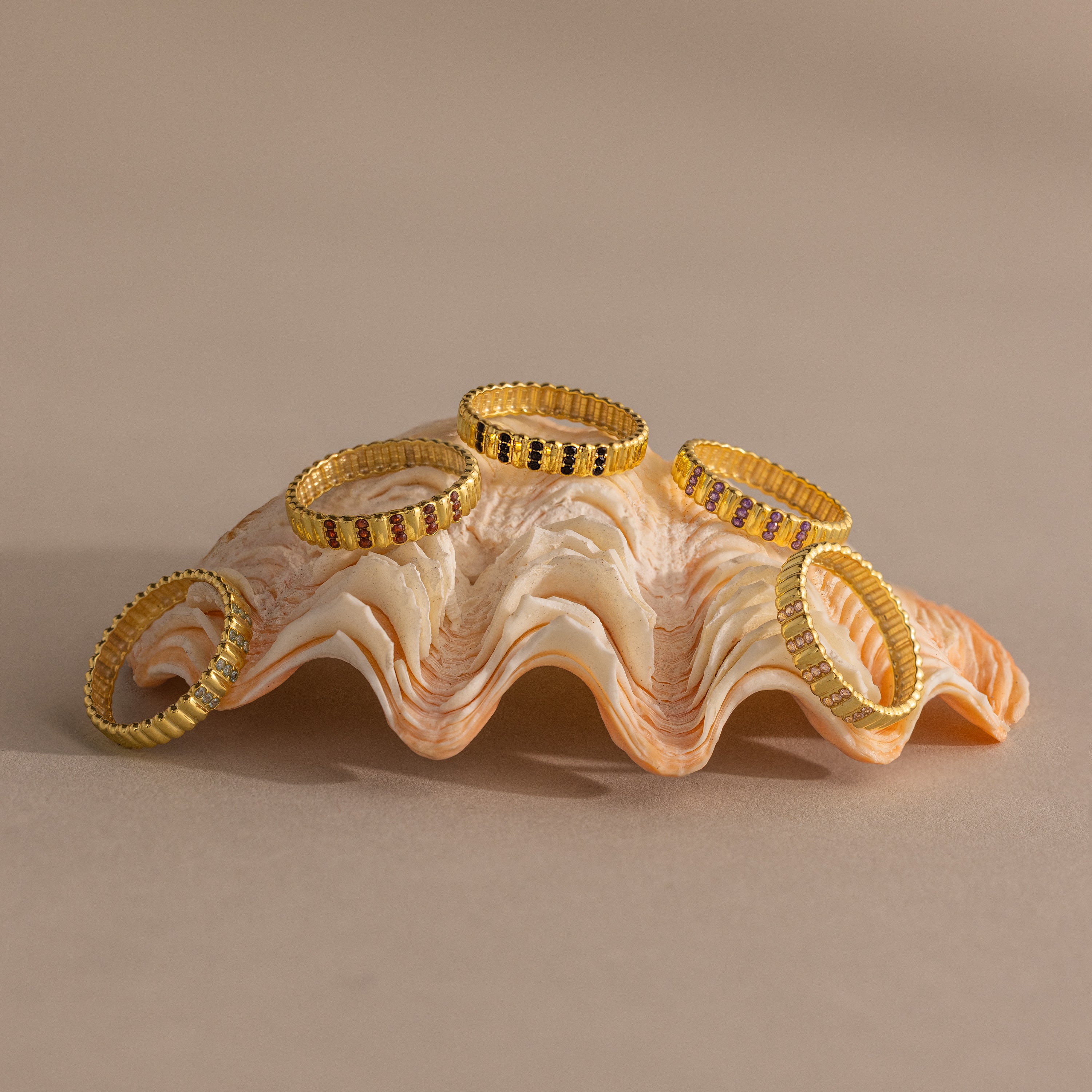 Five gold rings, including the Ribbed Triple Birthstone Ring, are adorned with colored gems and displayed on a seashell against a neutral beige background.