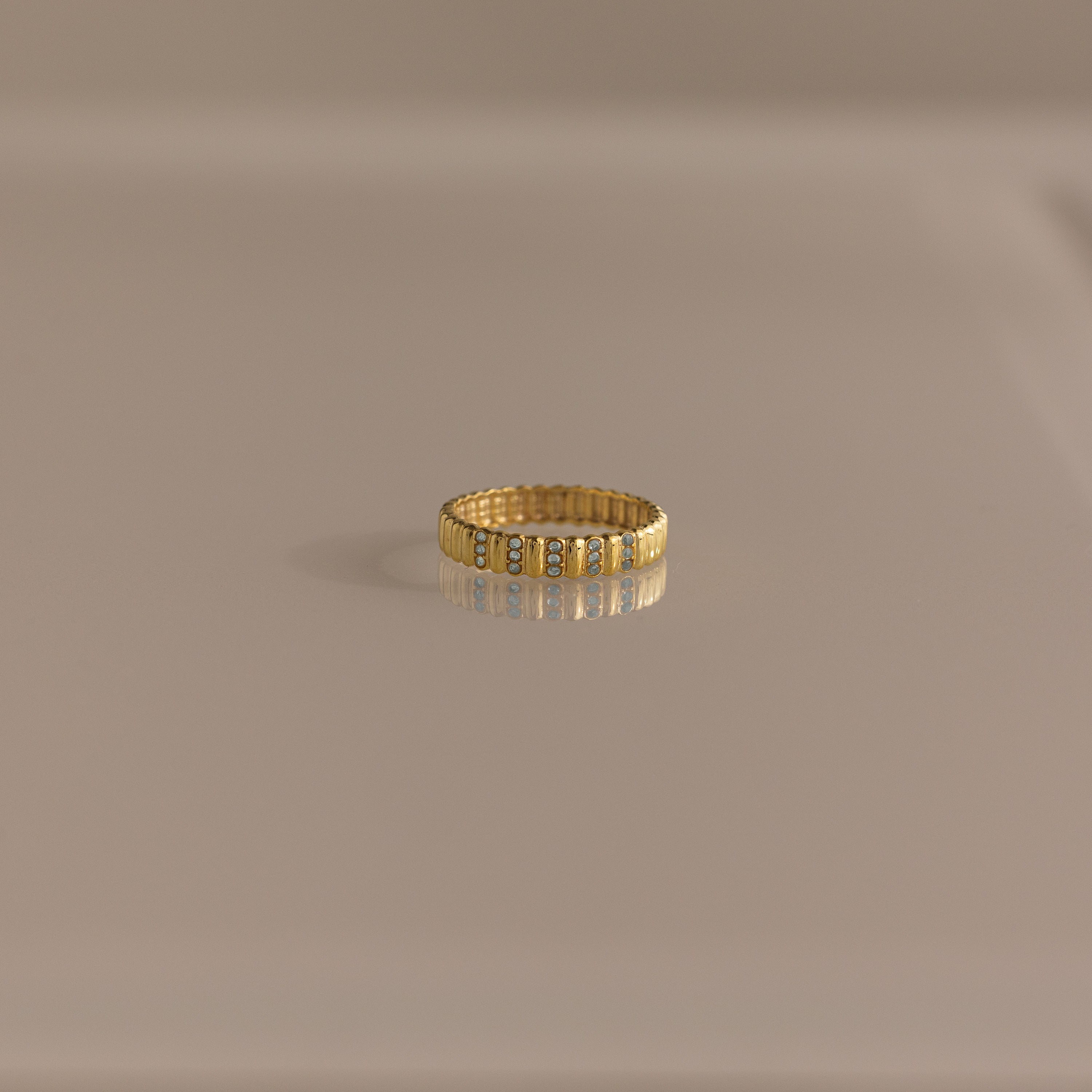 The Ribbed Triple Aquamarine Ring features a sculptural ridged band set with three clear aquamarine stones, glowing atop a smooth beige surface—perfect as a delicate birthstone ring.